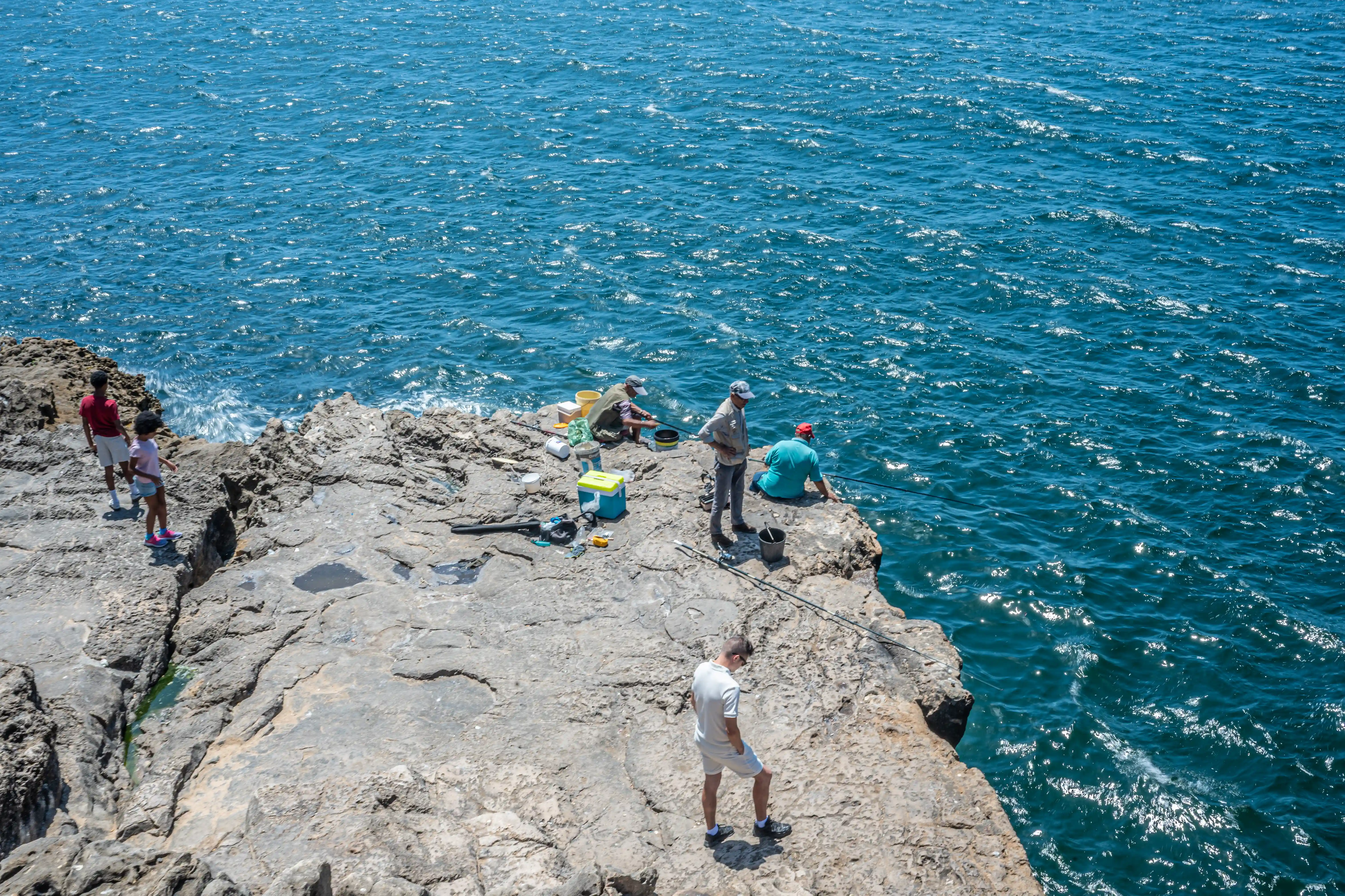 Several individuals are fishing from a sun-drenched, rocky coastline overlooking a blue, choppy sea. Two children stand on a higher part of the rocks, while four adults are spread out along the edge, some with fishing rods in hand.