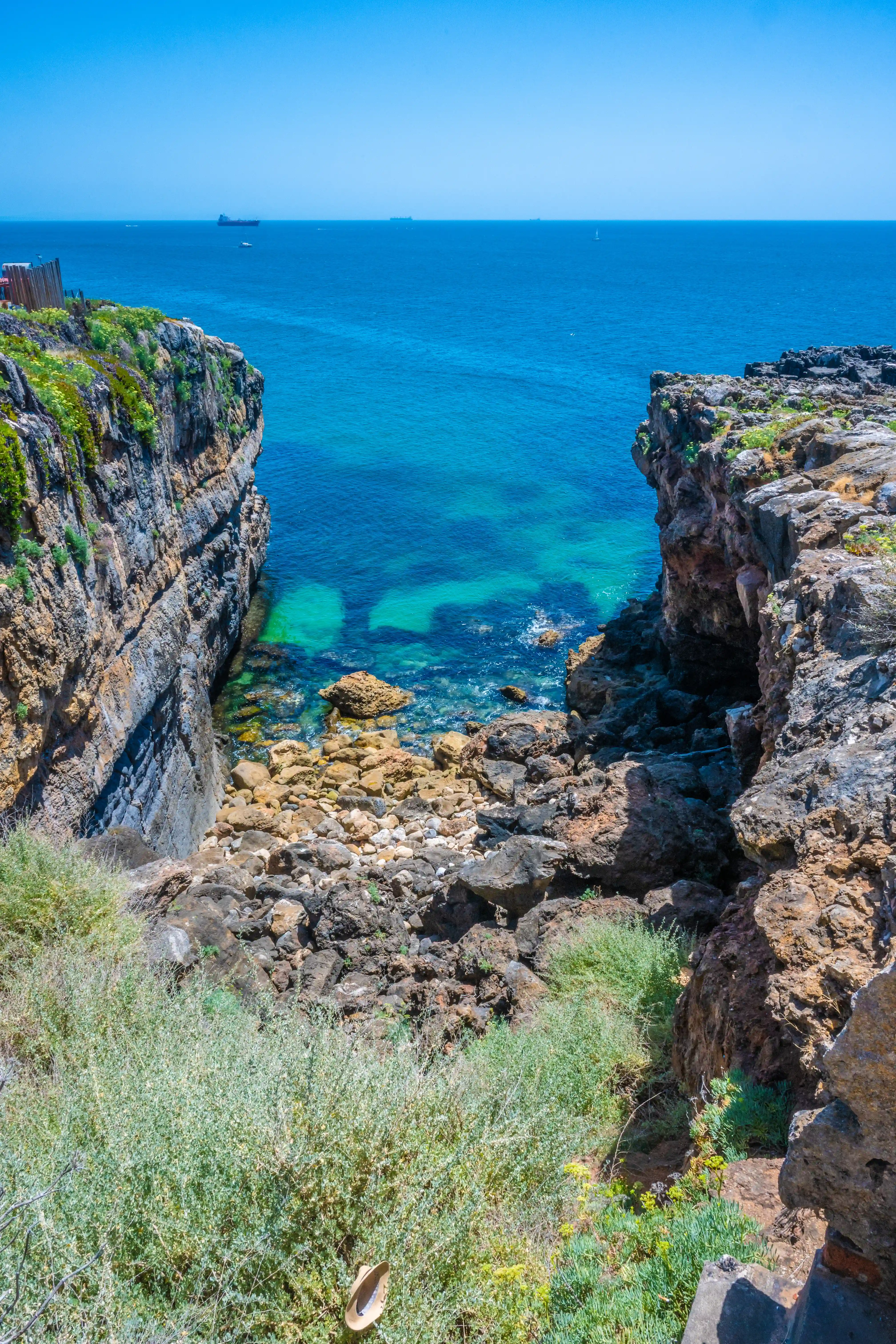 A high-angle view of a rugged coastline with steep, layered rock cliffs on either side, leading down to a calm, clear turquoise sea. Rocks and pebbles litter the small beach area at the base of the cliffs. A large cargo ship sails on the distant horizon under a clear blue sky.