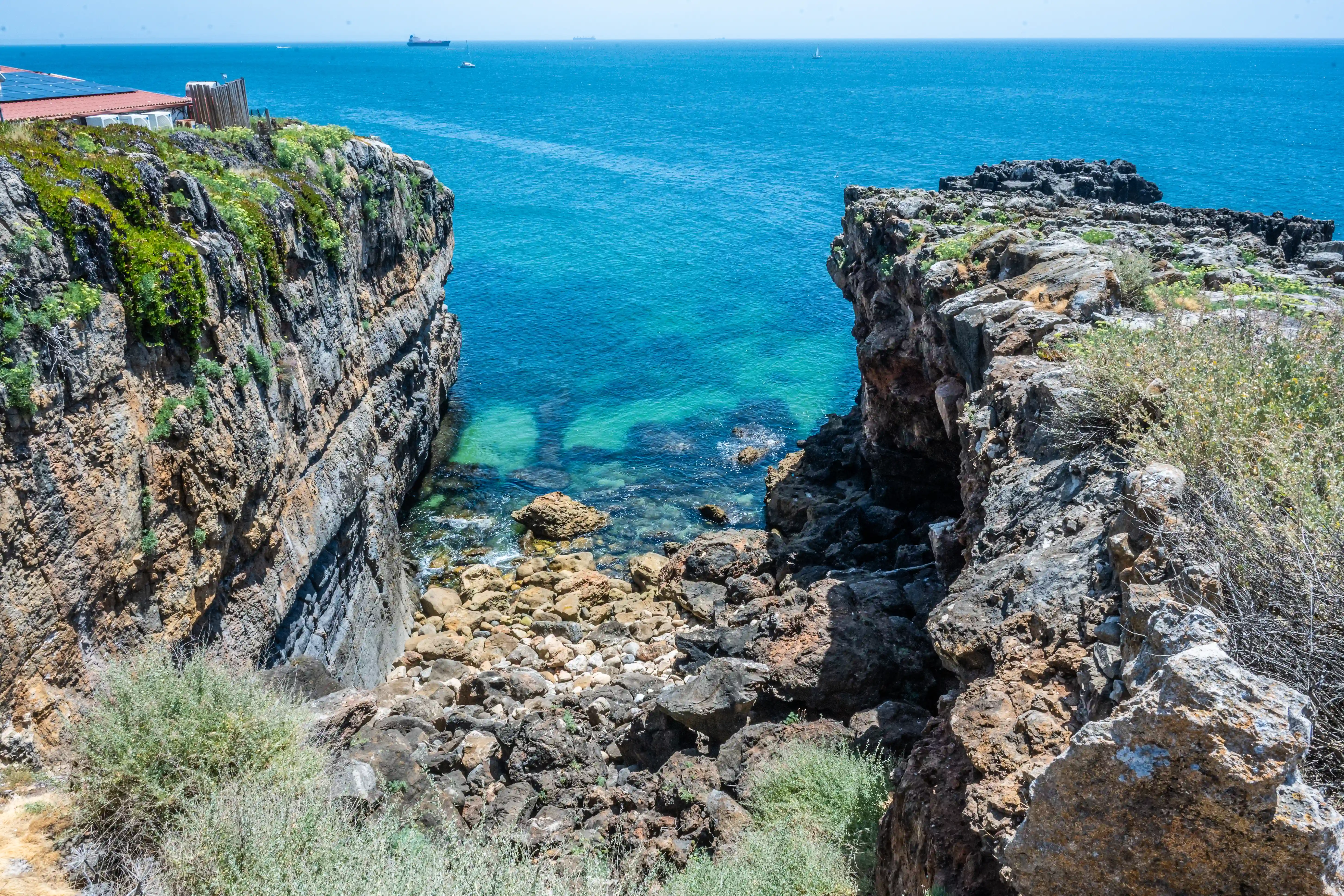 A view looking down into a rocky inlet where sheer, vegetated cliffs on either side meet the ocean. The water is a clear turquoise colour.