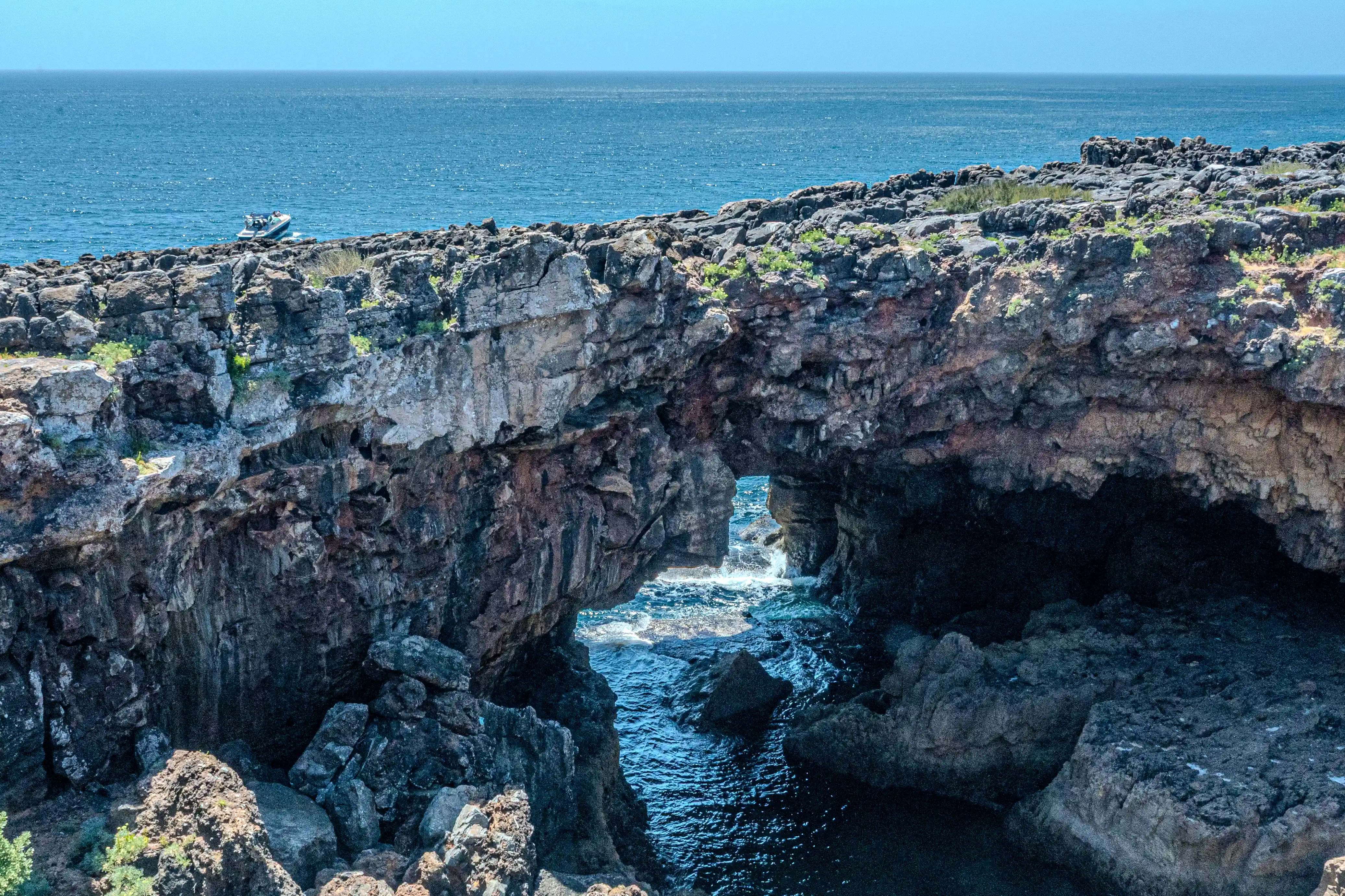 A natural rock arch formed by dark volcanic rocks, framing a small inlet where waves crash against the shore. The deep blue sea extends to the horizon under a clear sky, with a small boat visible in the distance