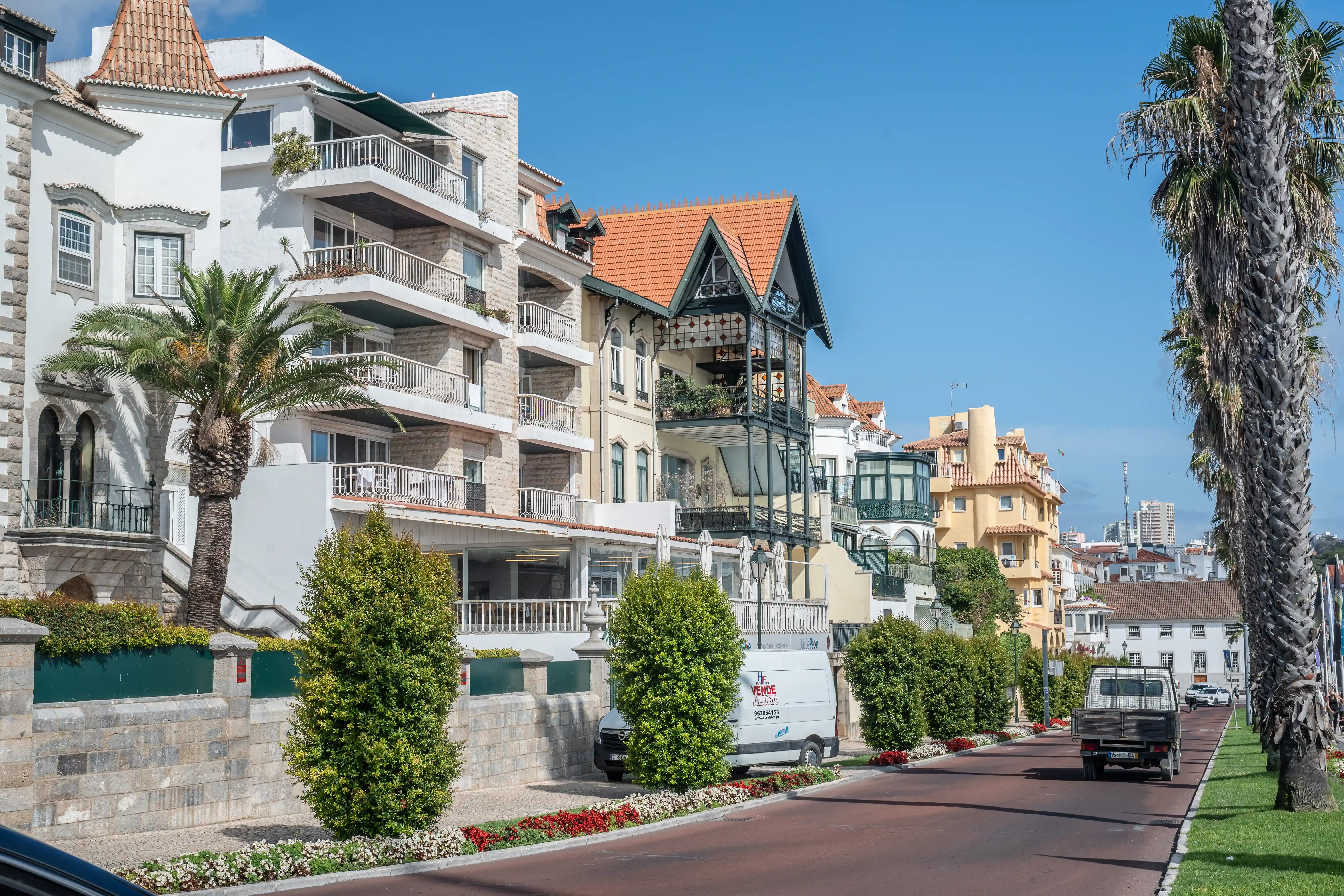 A street view with a line of white and pastel-coloured buildings, featuring balconies, windows, and pitched roofs, under a clear blue sky. A tall palm tree stands to the right, and a white van and a truck drive along the road.