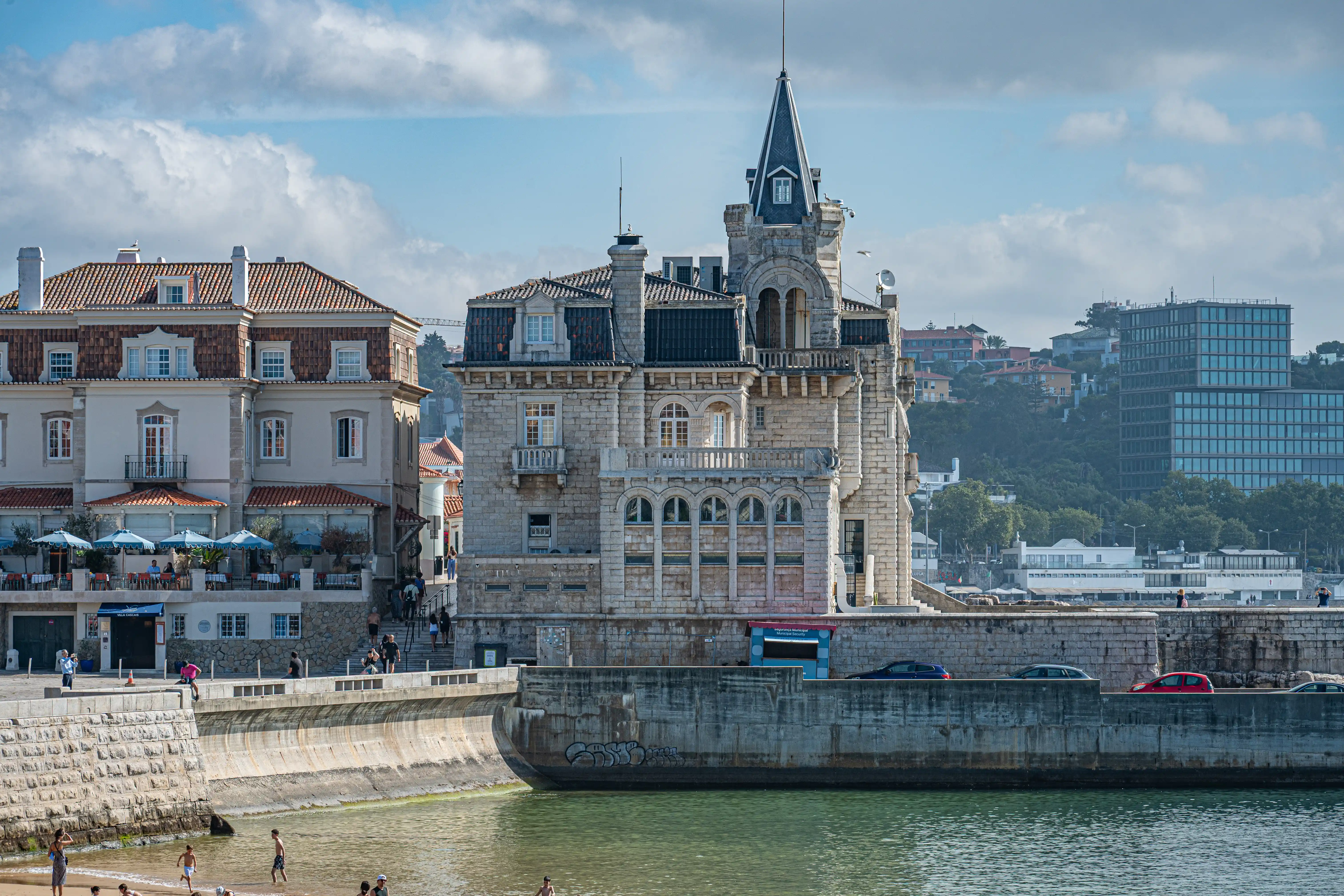 A stone mansion with a pointed turret overlooks a beach where people are swimming and sunbathing. A promenade with cars is in the mid-ground, and a modern glass building is visible in the distance on a hill.