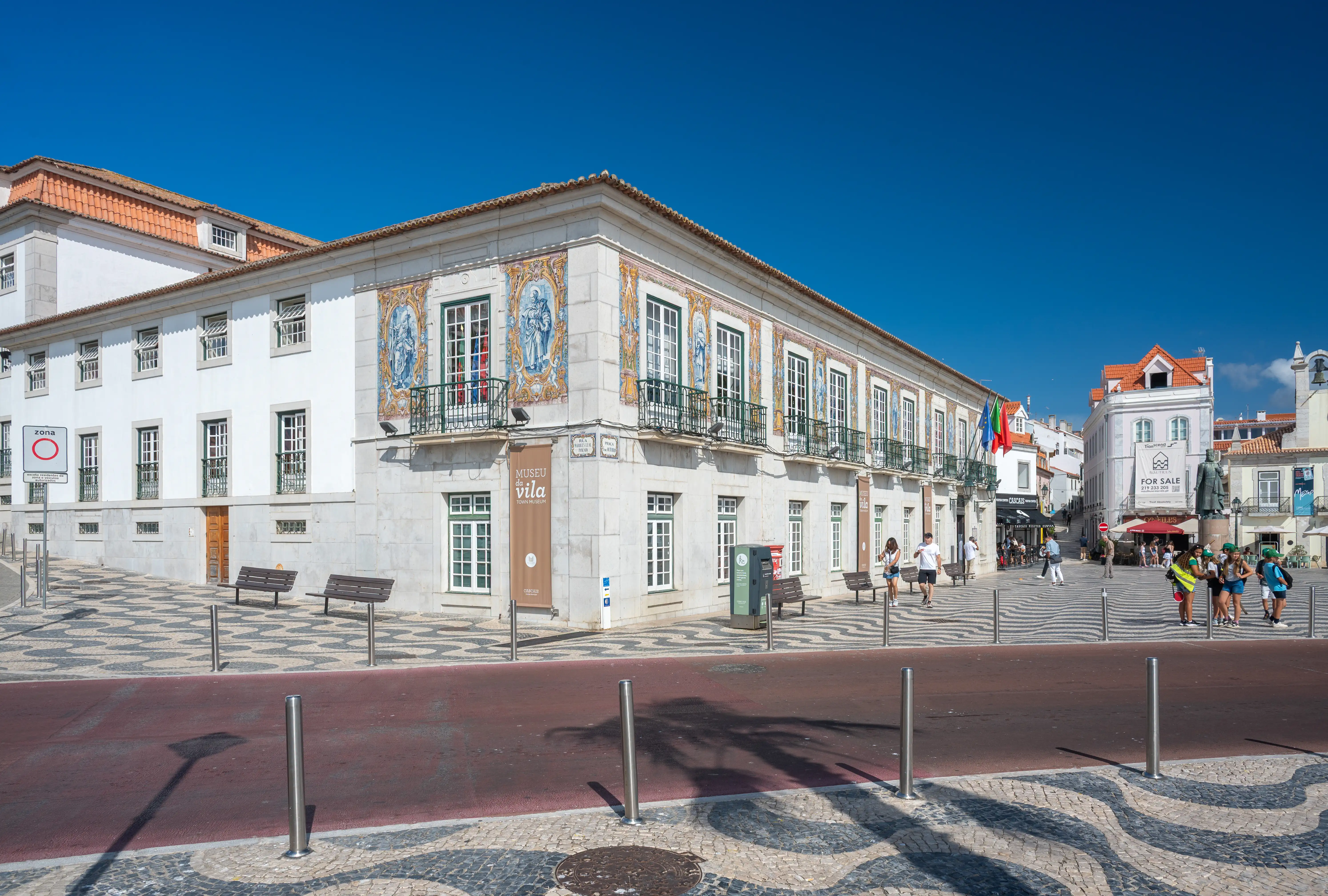 The exterior of the Museu da Vila, a white building with decorative blue and white azulejo tiles, stands in a town square paved with a black and white wavy pattern. Several people are walking in the background