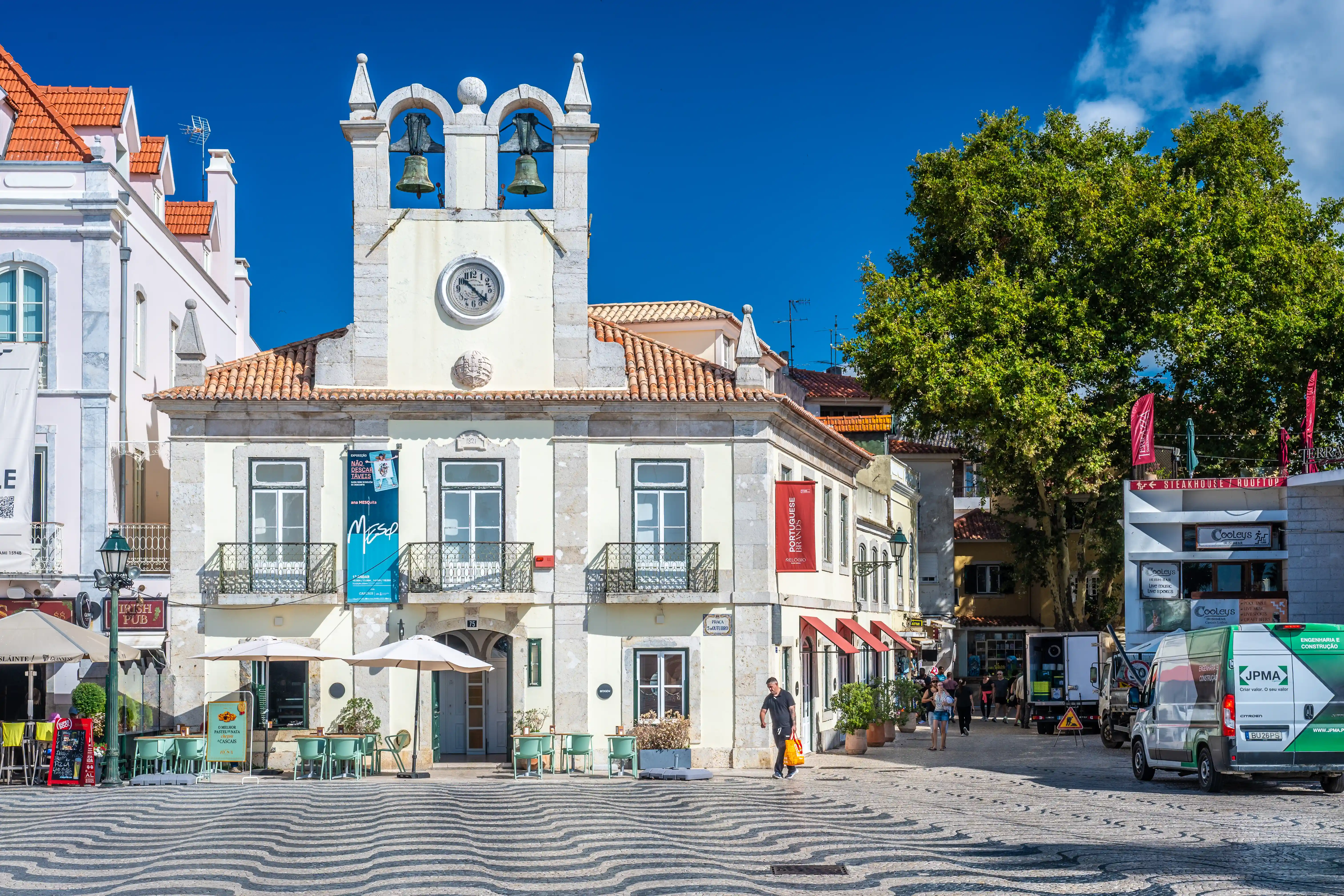 A man walks across a patterned pavement in a European town square towards a pale yellow building topped with a clock tower and two bells. Outdoor cafe tables and umbrellas are scattered across the square, and a large green tree provides shade on the right side of the image.