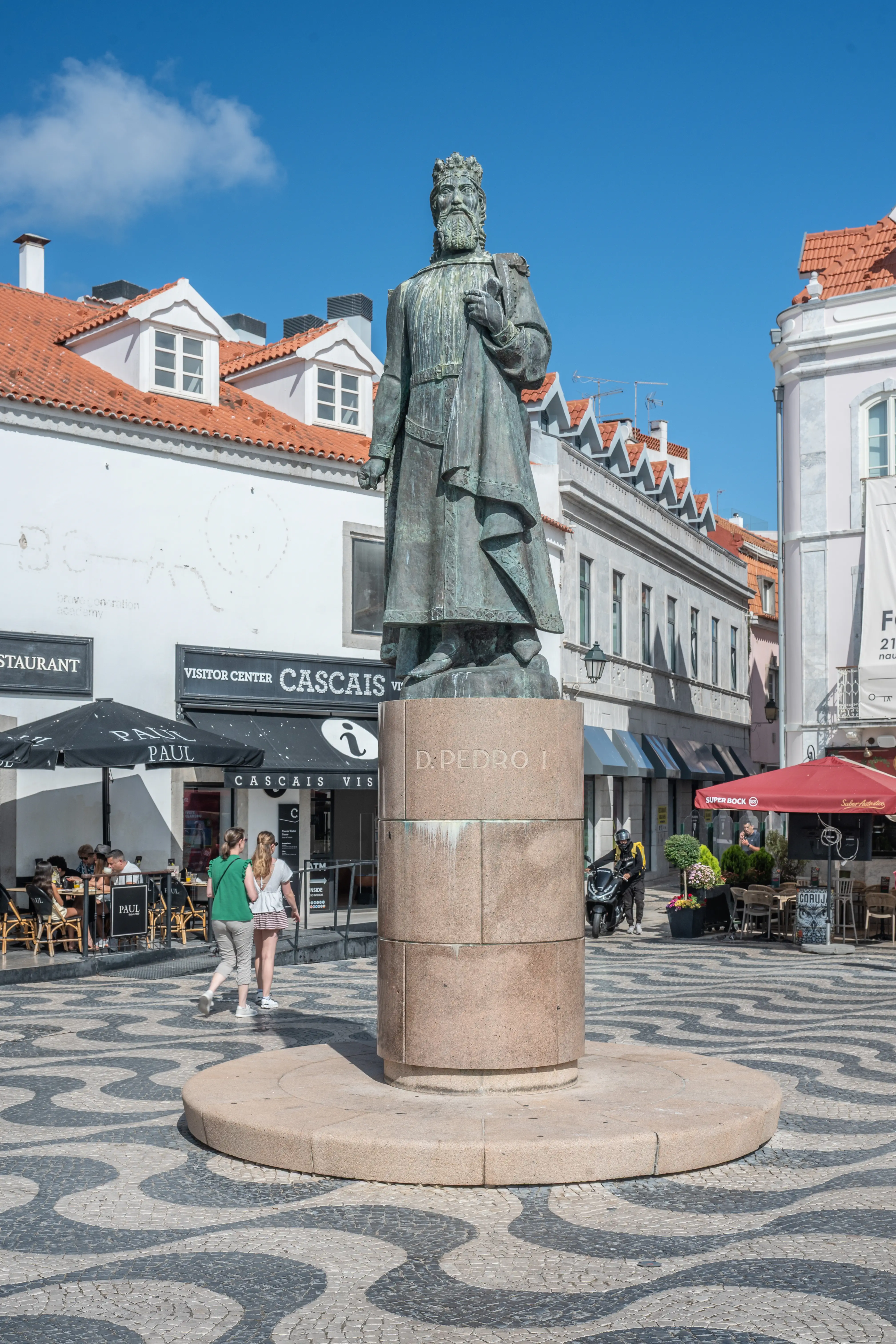 A bronze statue of a historical figure stands on a cylindrical stone pedestal inscribed with 'D. PEDRO I' in a bustling square. The square is paved with black-and-white wave-patterned tiles. Buildings with outdoor seating surround the square, and people are gathered around. The sky is clear and blue.