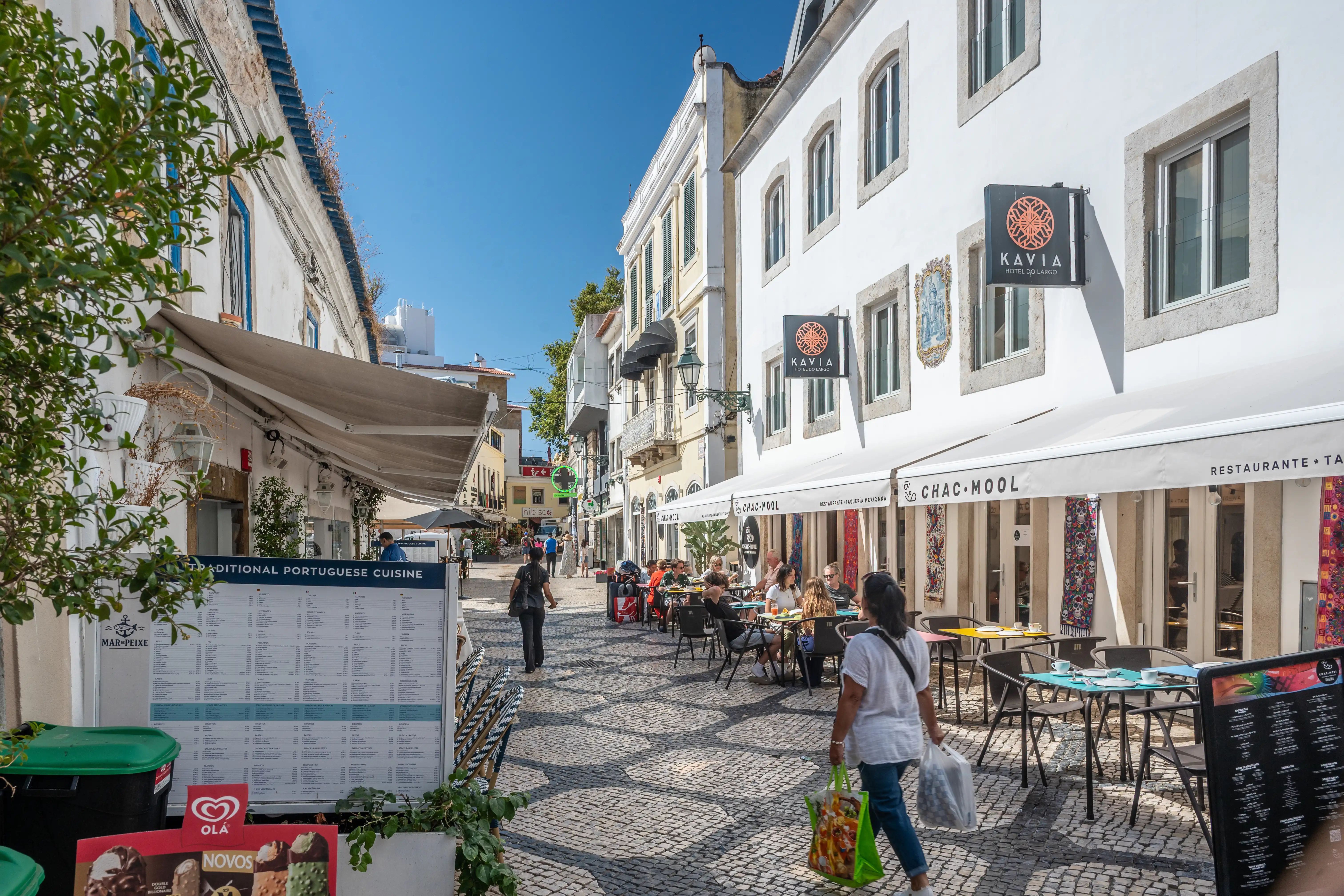 A sunny street in a Portuguese town lined with white buildings and outdoor cafes. People are seated at tables, and a woman walks down the cobblestone street.
