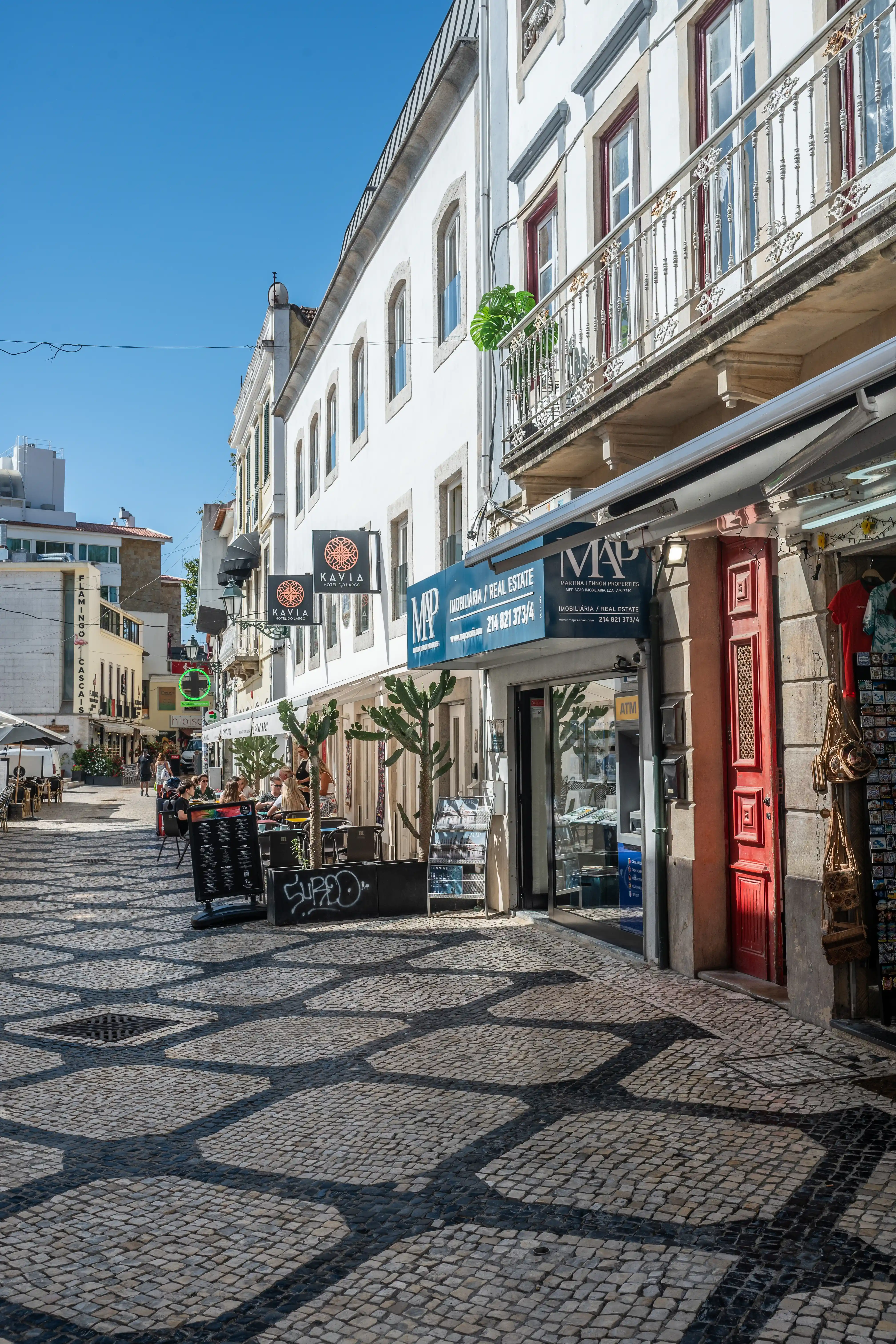 A sun-drenched pedestrian street lined with white buildings and outdoor cafes. A real estate agency with a blue sign is visible, alongside traditional cobblestone paving and shadows cast by buildings and trees.