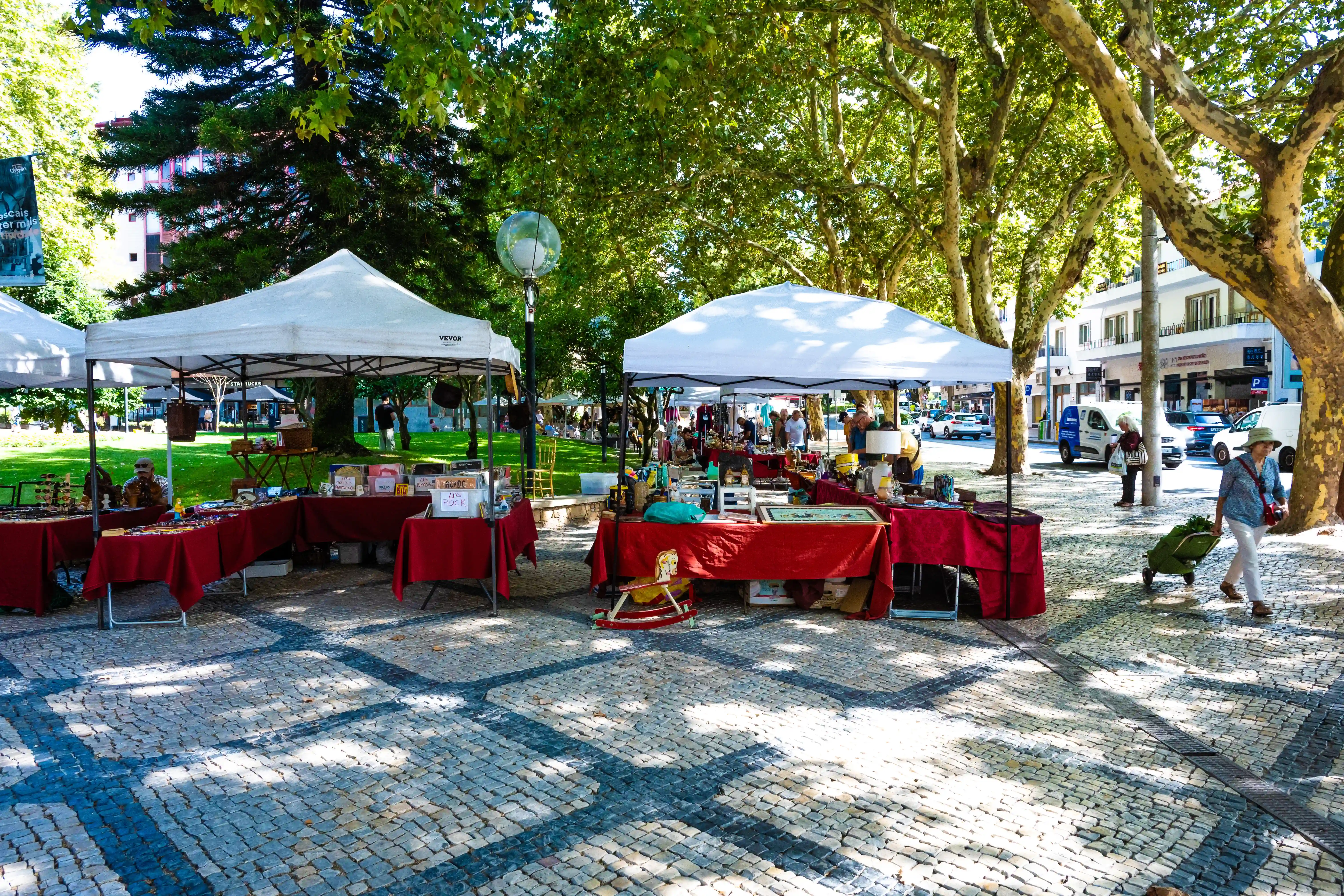 An outdoor market with stalls under white canopies displays various items for sale. People are browsing, and a woman is walking with a wheeled shopping trolley. The ground is paved with cobbles and shaded by trees.