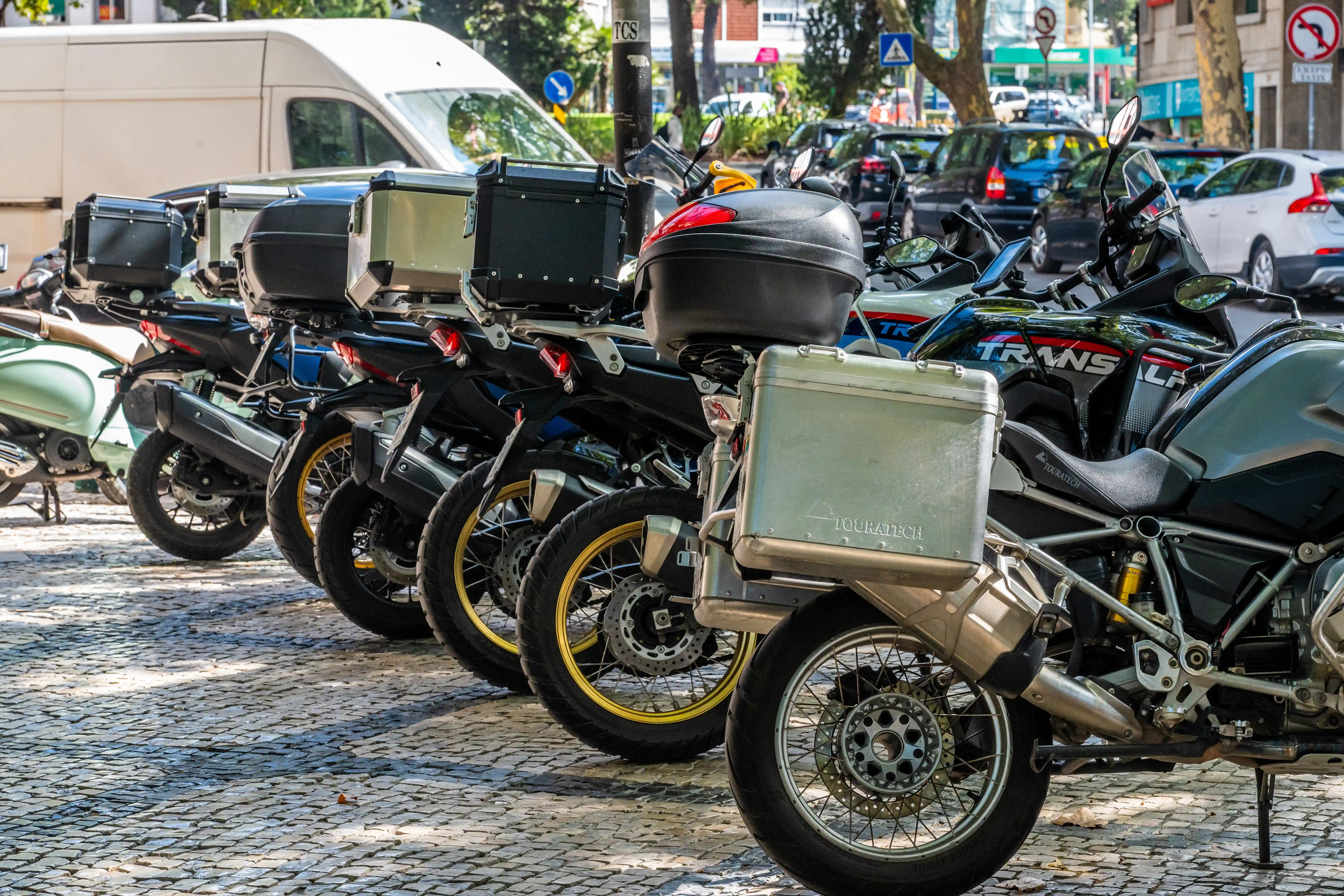 Several adventure motorcycles are parked in a row on a cobblestone street. The motorcycles have large storage boxes attached to the rear, with one box labelled &laquo;Touratech&raquo;.
