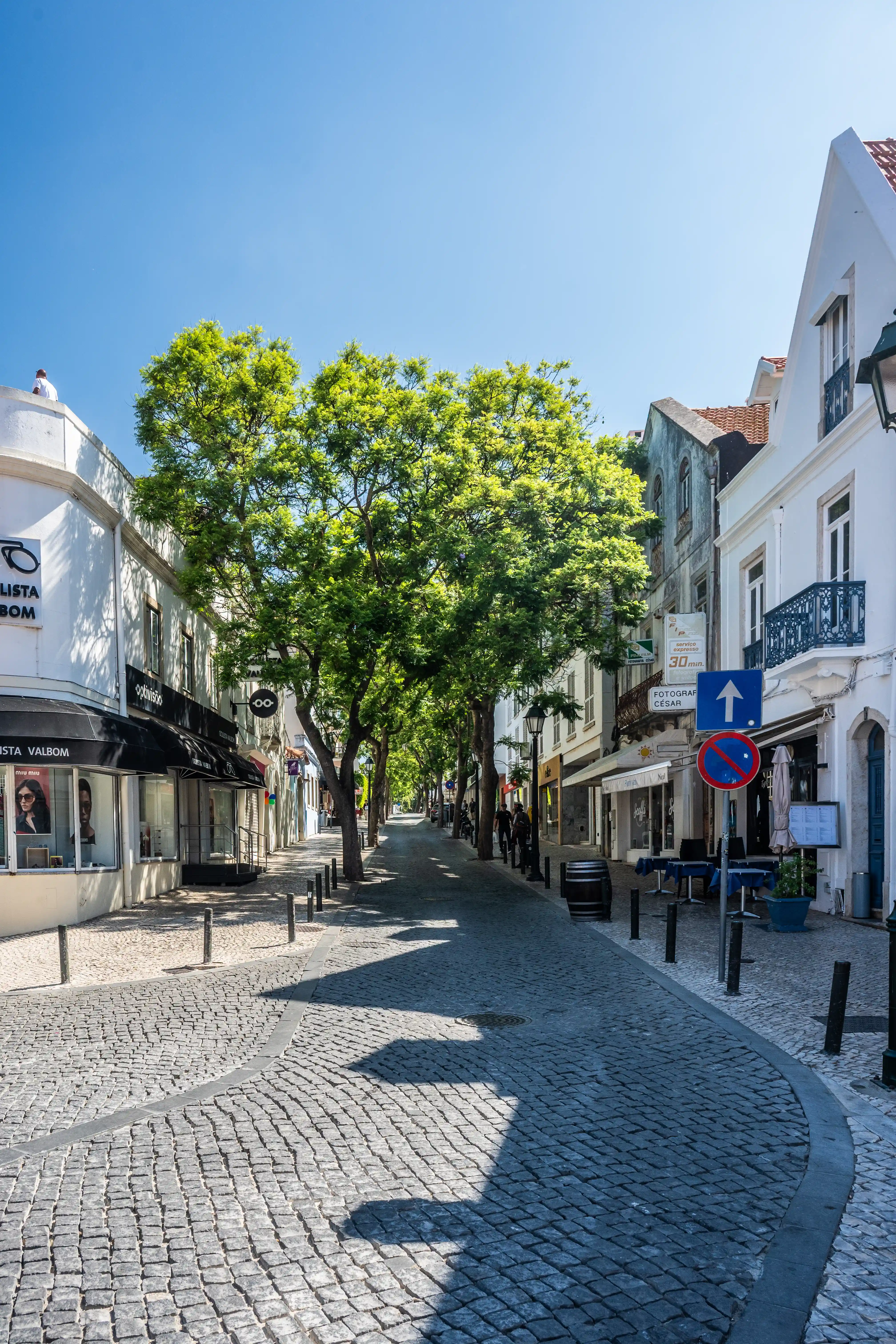 A sun-drenched pedestrian street lined with white buildings and outdoor cafes. A real estate agency with a blue sign is visible, alongside traditional cobblestone paving and shadows cast by buildings and trees.