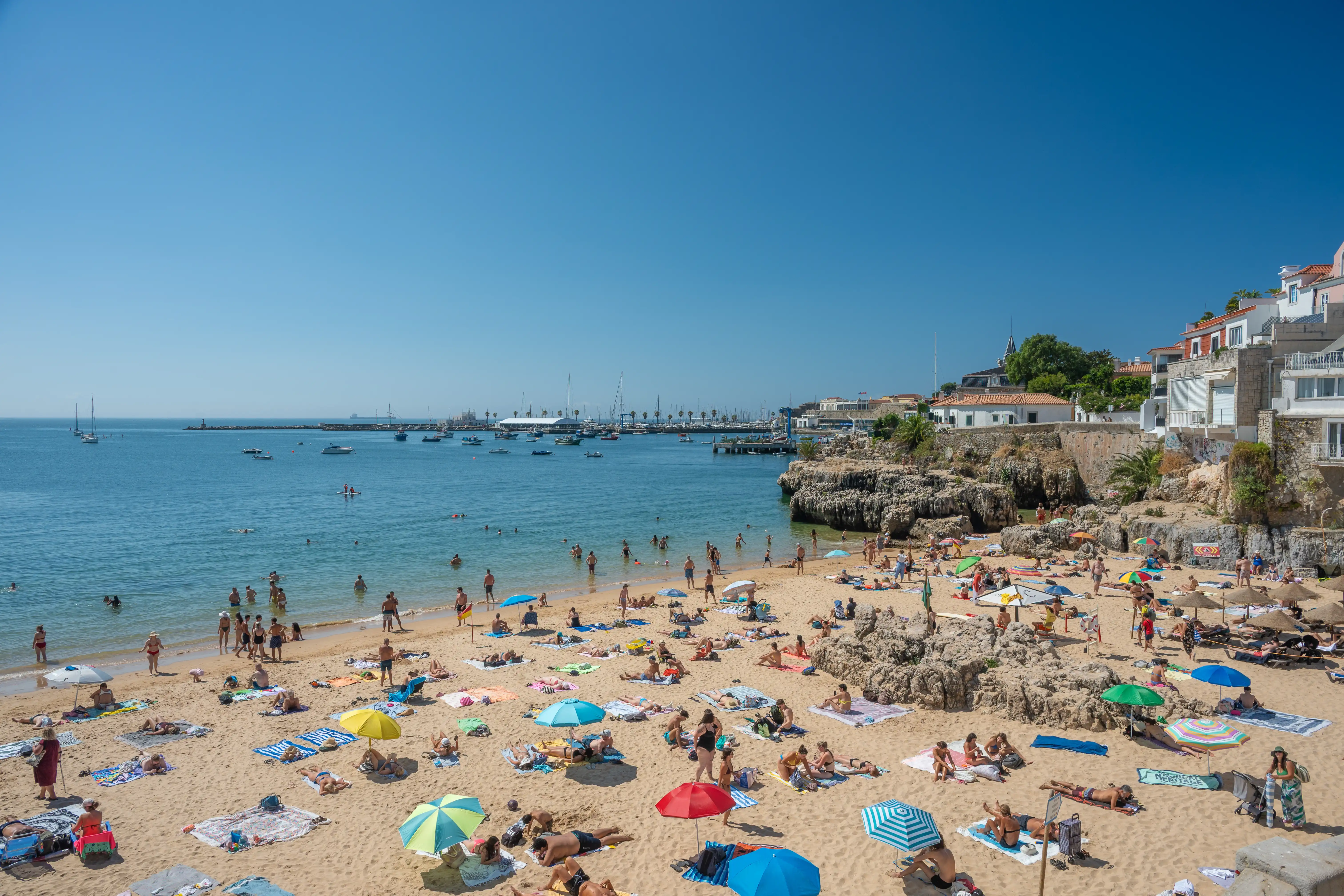 A high-angle view of a crowded beach with people sunbathing on the sand and swimming in the ocean. Buildings and a harbour with boats are visible on the coastline under a bright blue sky.
