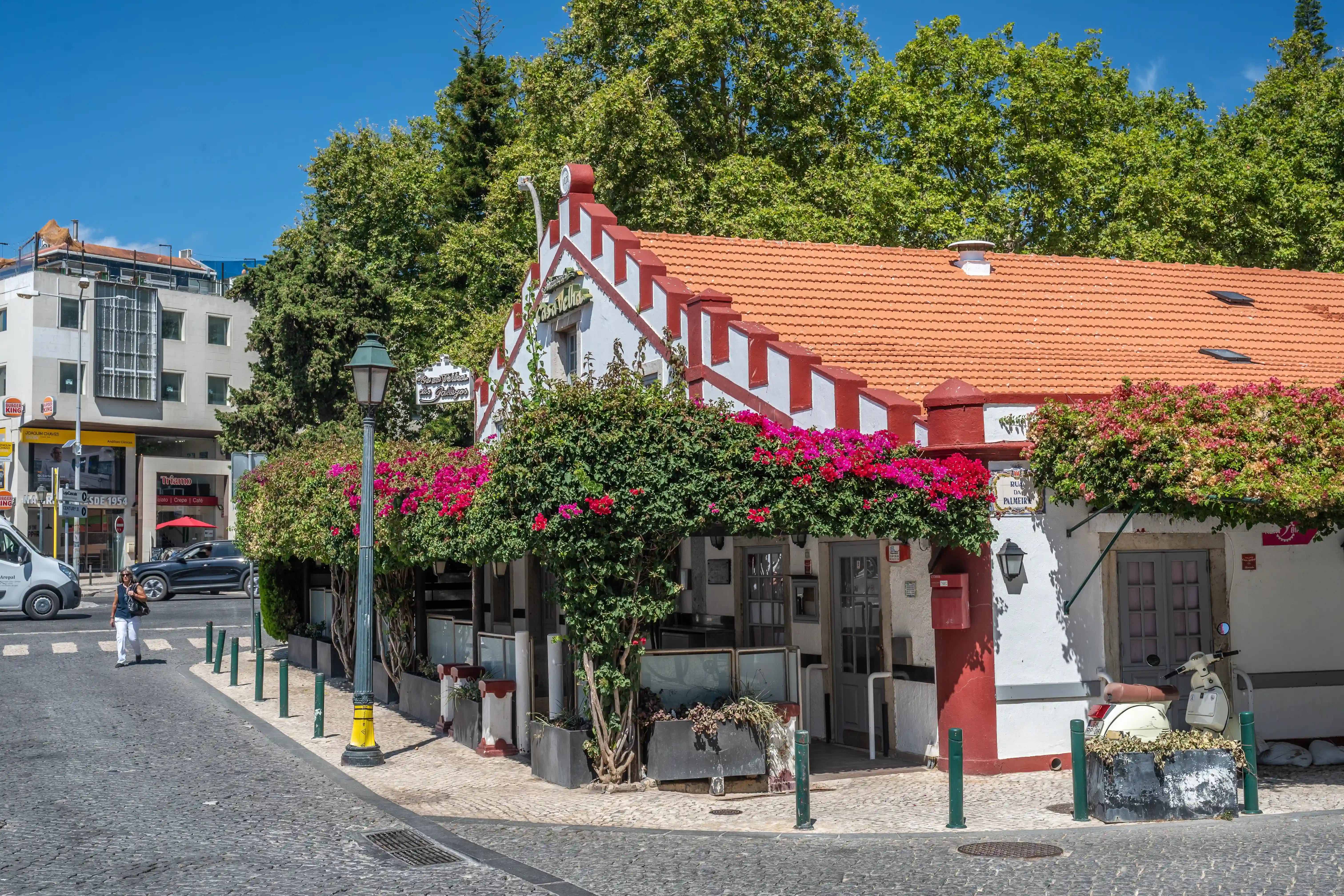 A quaint white building with a red-tiled roof and decorative red trim, draped in lush pink bougainvillea, stands beside a cobblestone street. A vintage white scooter is parked in front.