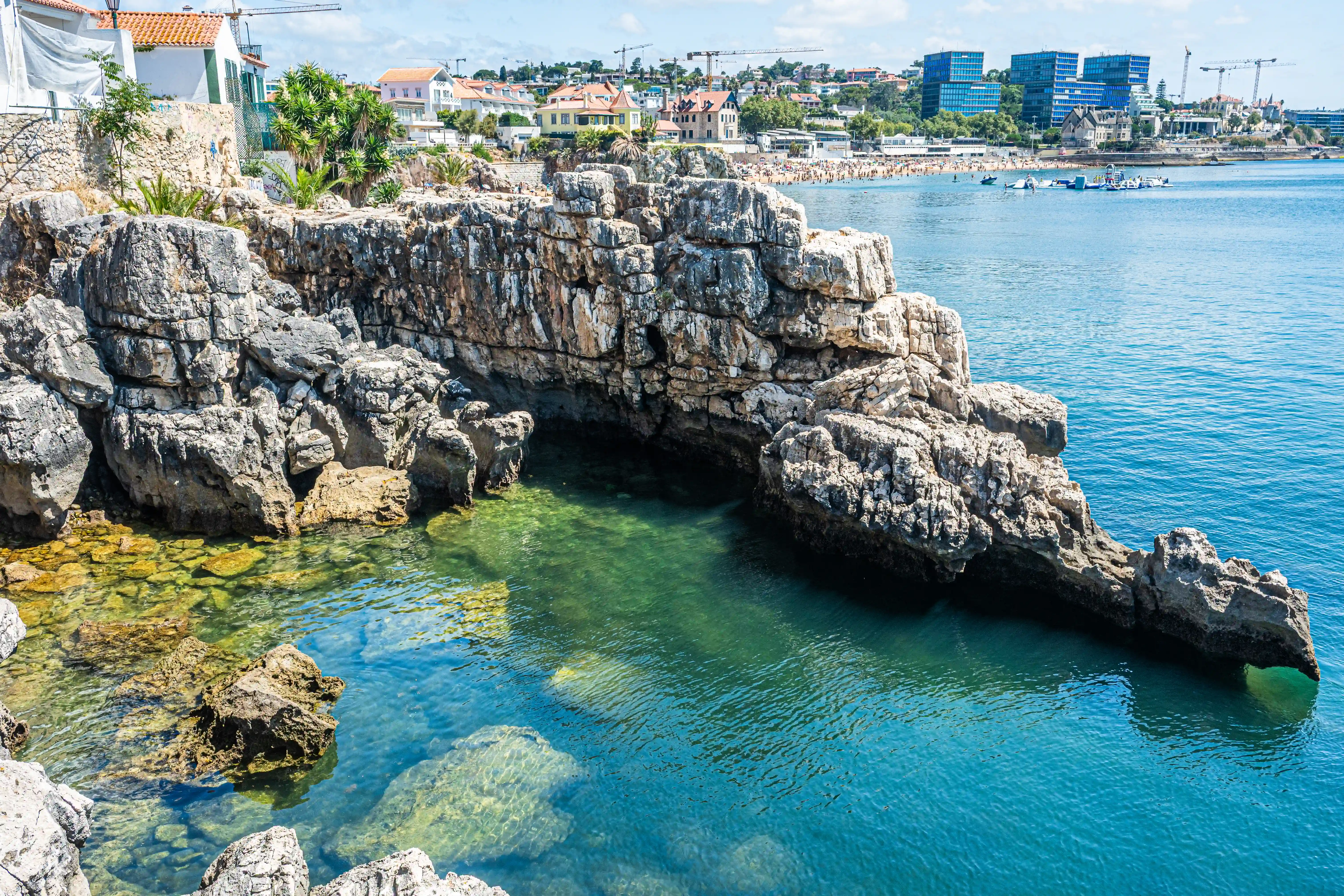 A rugged coastal cliff with jagged rocks and clear turquoise waters in the foreground. In the background, modern buildings and construction cranes rise against the horizon. A crowded beach with people is visible between the cliff and the city.