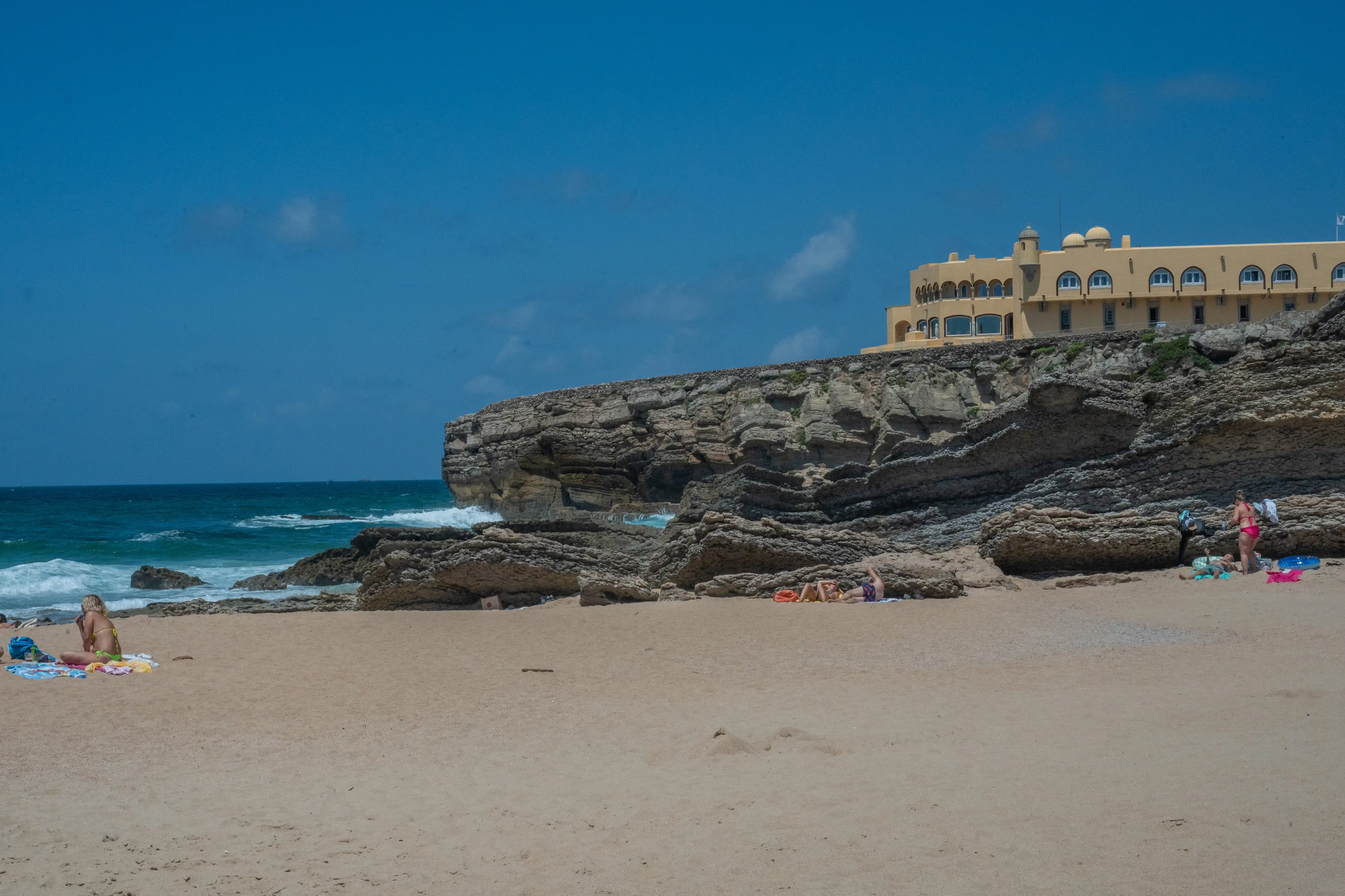  A sandy beach with several sunbathers in the foreground, leading to rocky cliffs topped by a large, light-coloured hotel. The blue sea with small waves is visible to the left, under a clear blue sky.