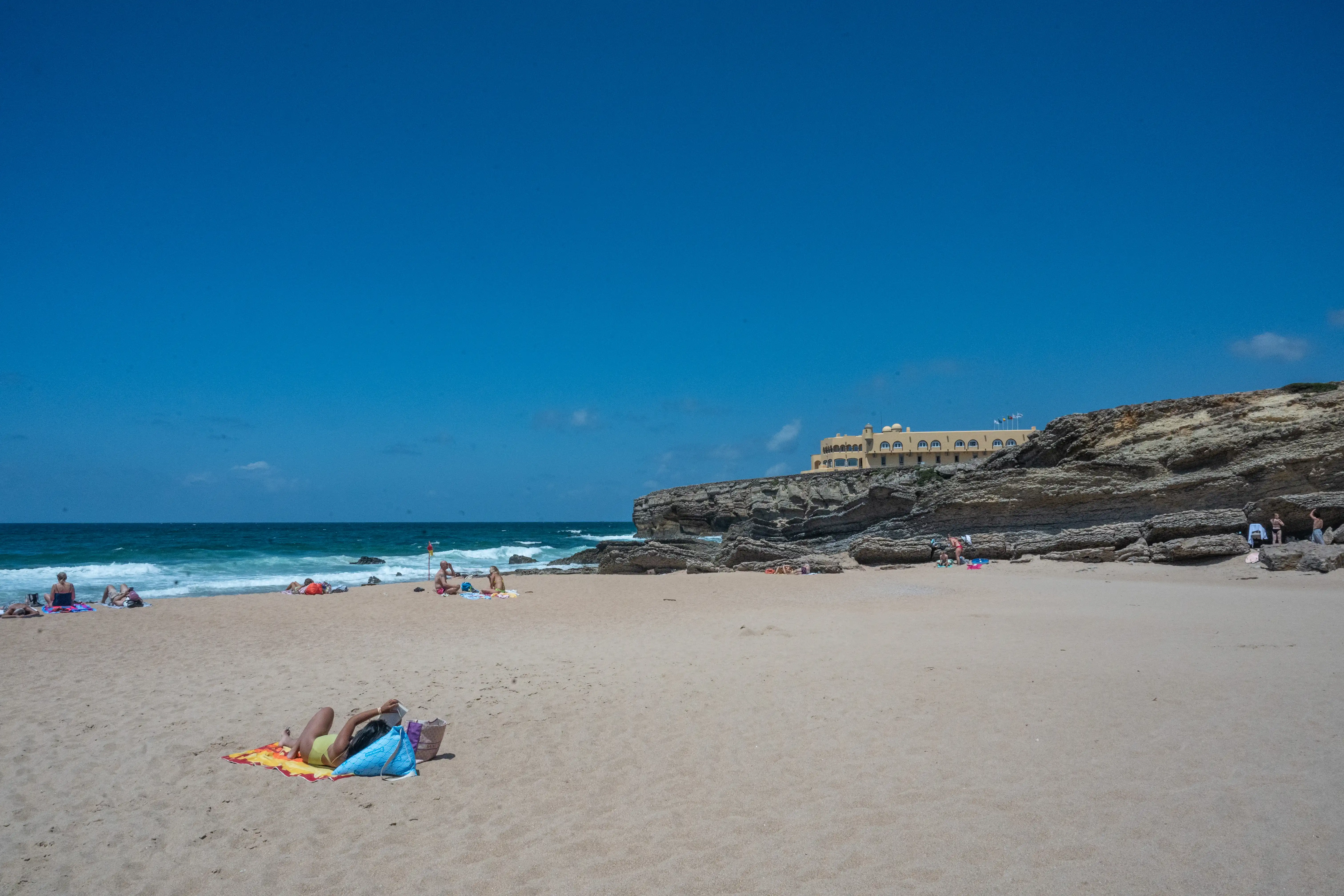 A serene beach scene with people relaxing on towels under a clear blue sky. The turquoise sea gently laps against the rocks, and a yellow building with arched windows and flags sits on a rocky cliff in the background