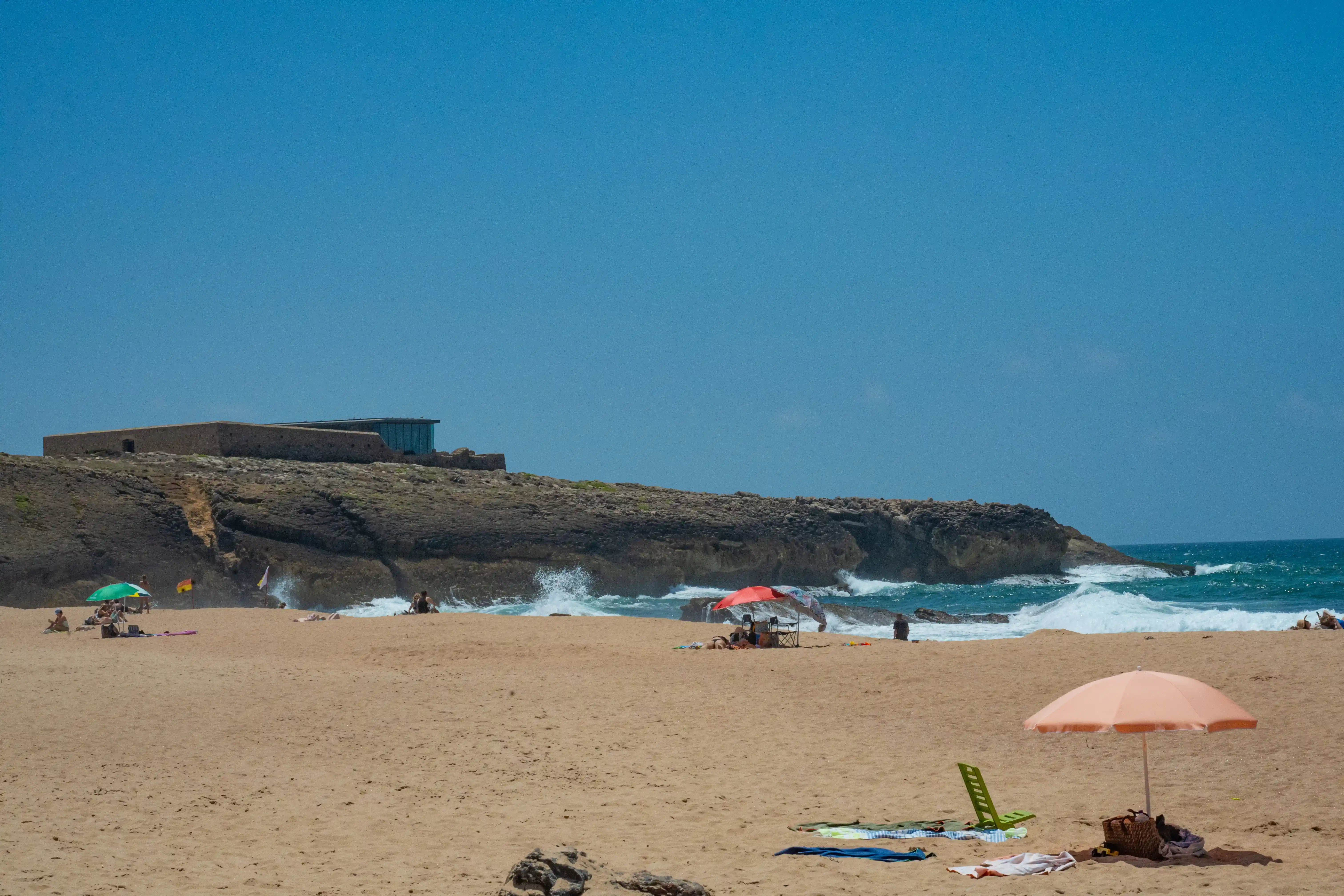A wide shot of a sandy beach on a sunny day, with a building on a cliff in the background and waves breaking on the shore.