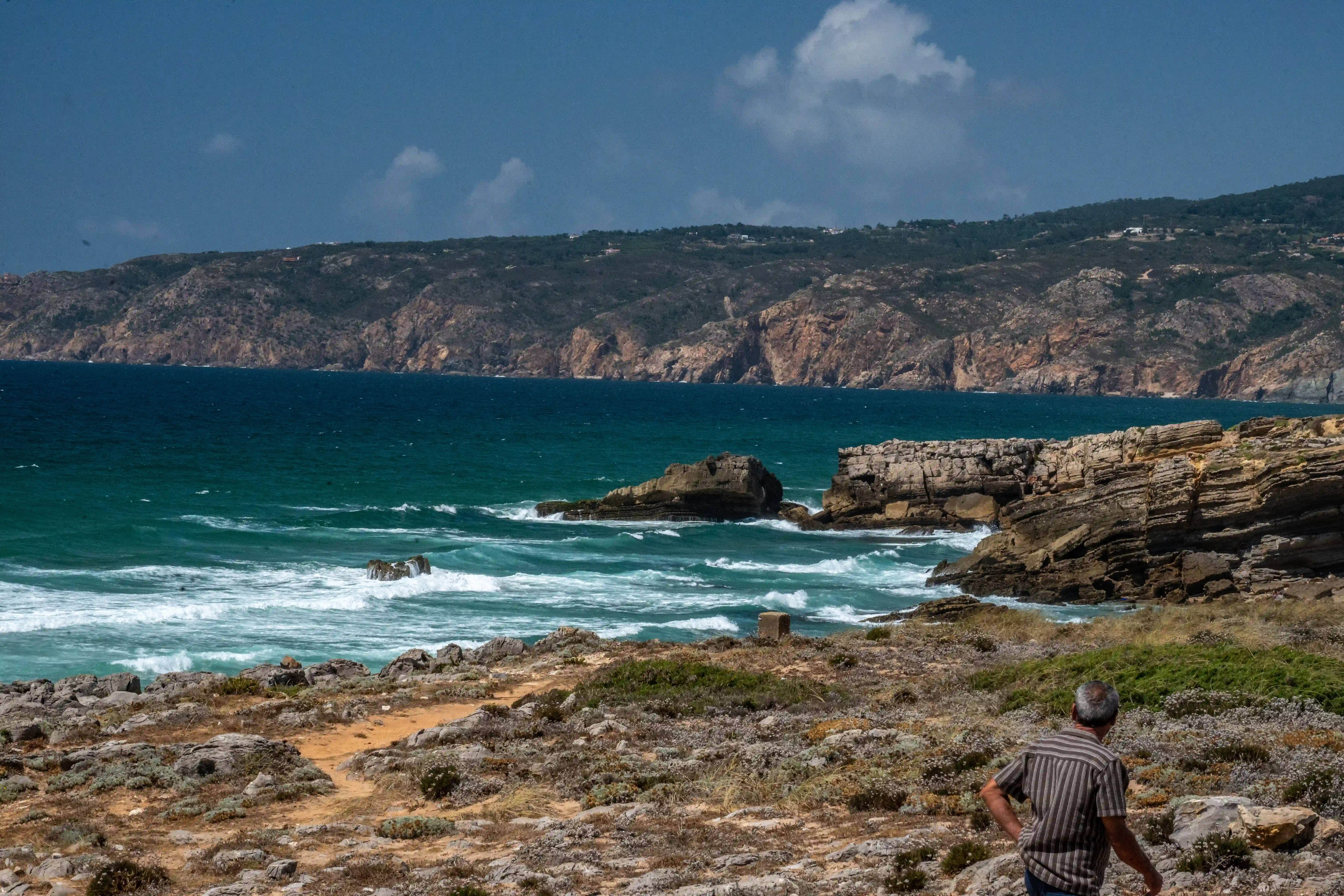 A man in a striped shirt stands on a rocky coastal path, looking out at the deep blue sea. Waves crash against the rugged cliffs, and the sky is overcast