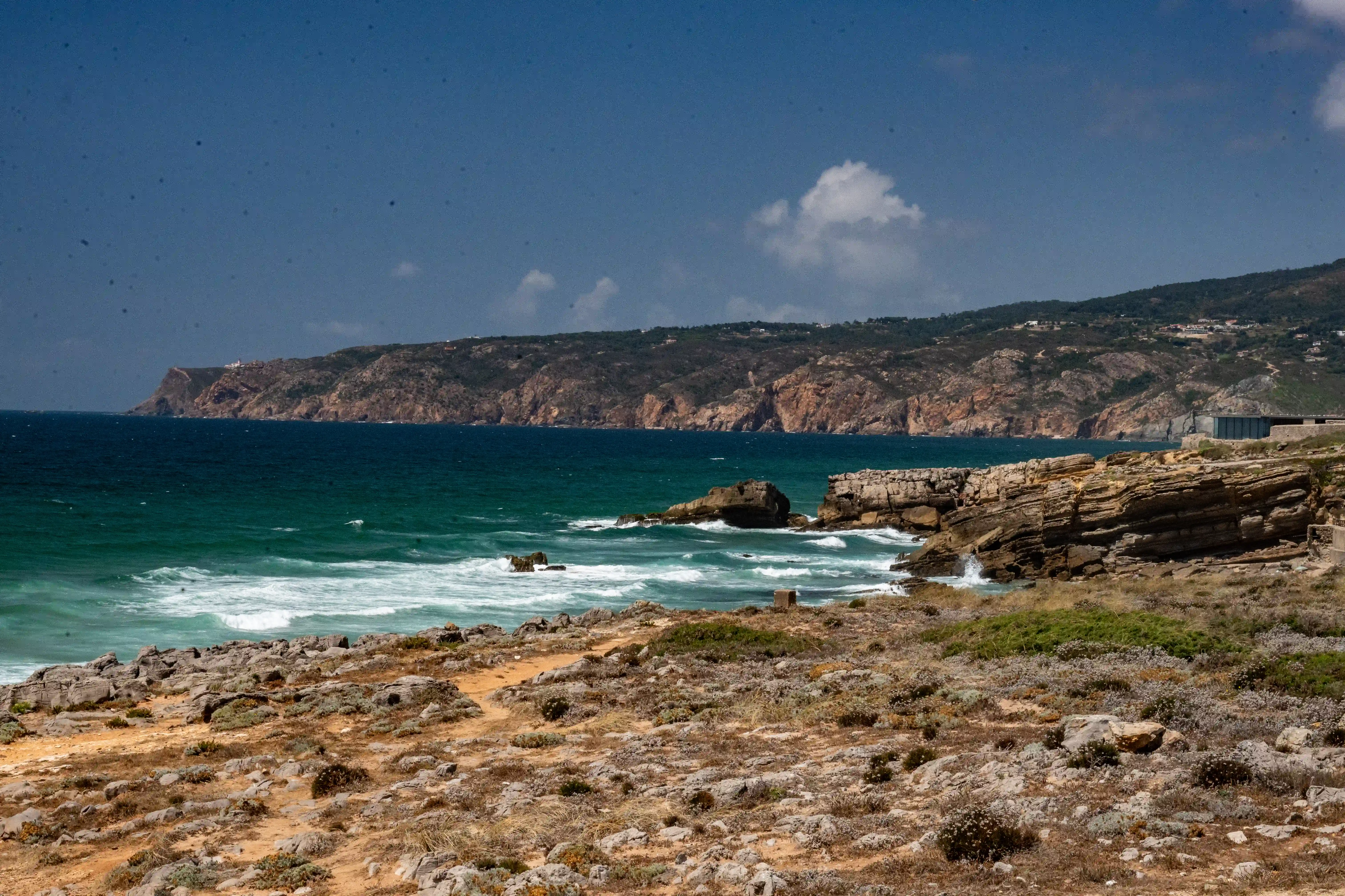 A man in a striped shirt stands on a rocky coastal path, looking out at the deep blue sea. Waves crash against the rugged cliffs, and the sky is overcast