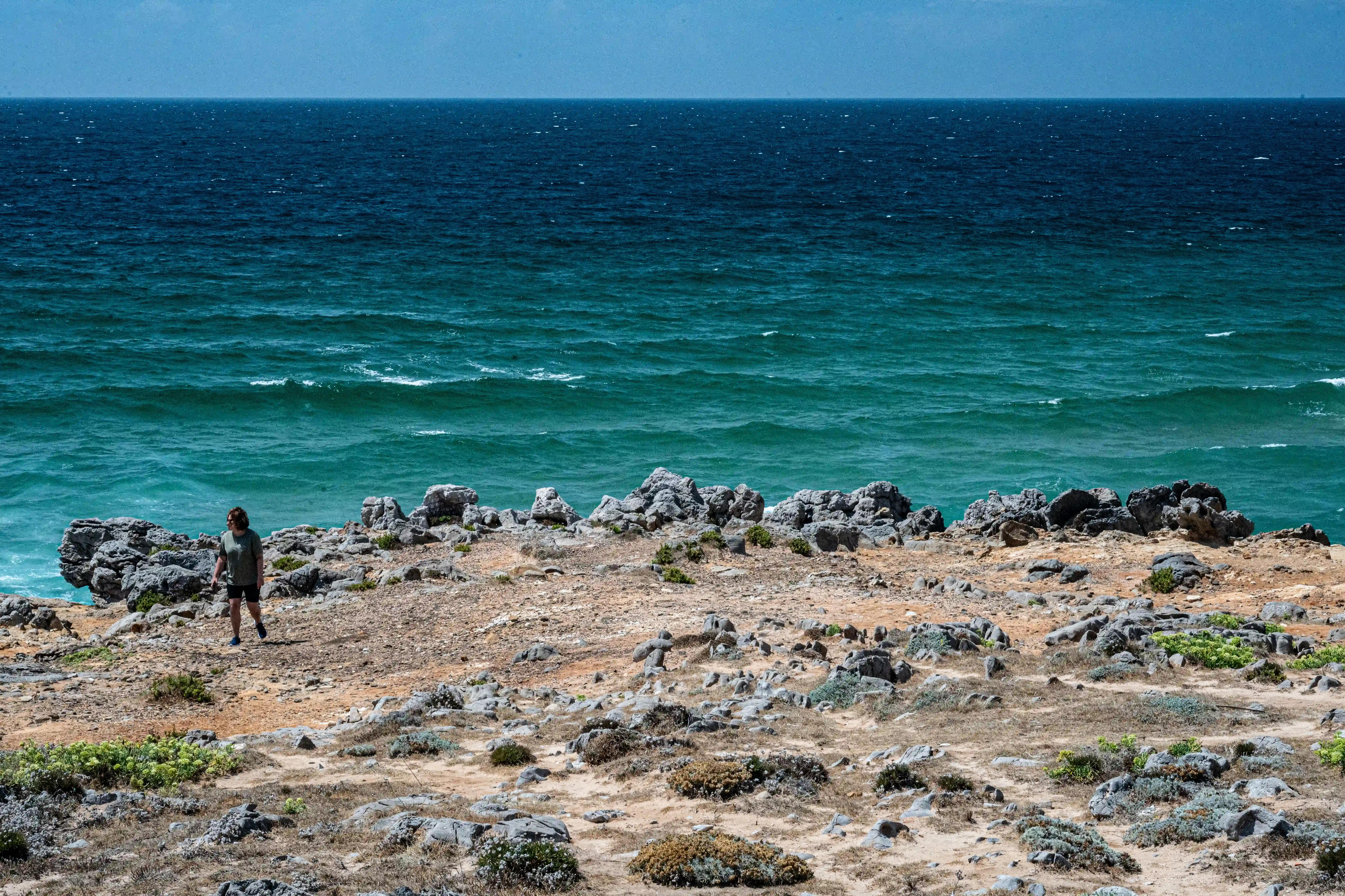 A person walks on a rocky, dry terrain overlooking a vast expanse of dark turquoise ocean.