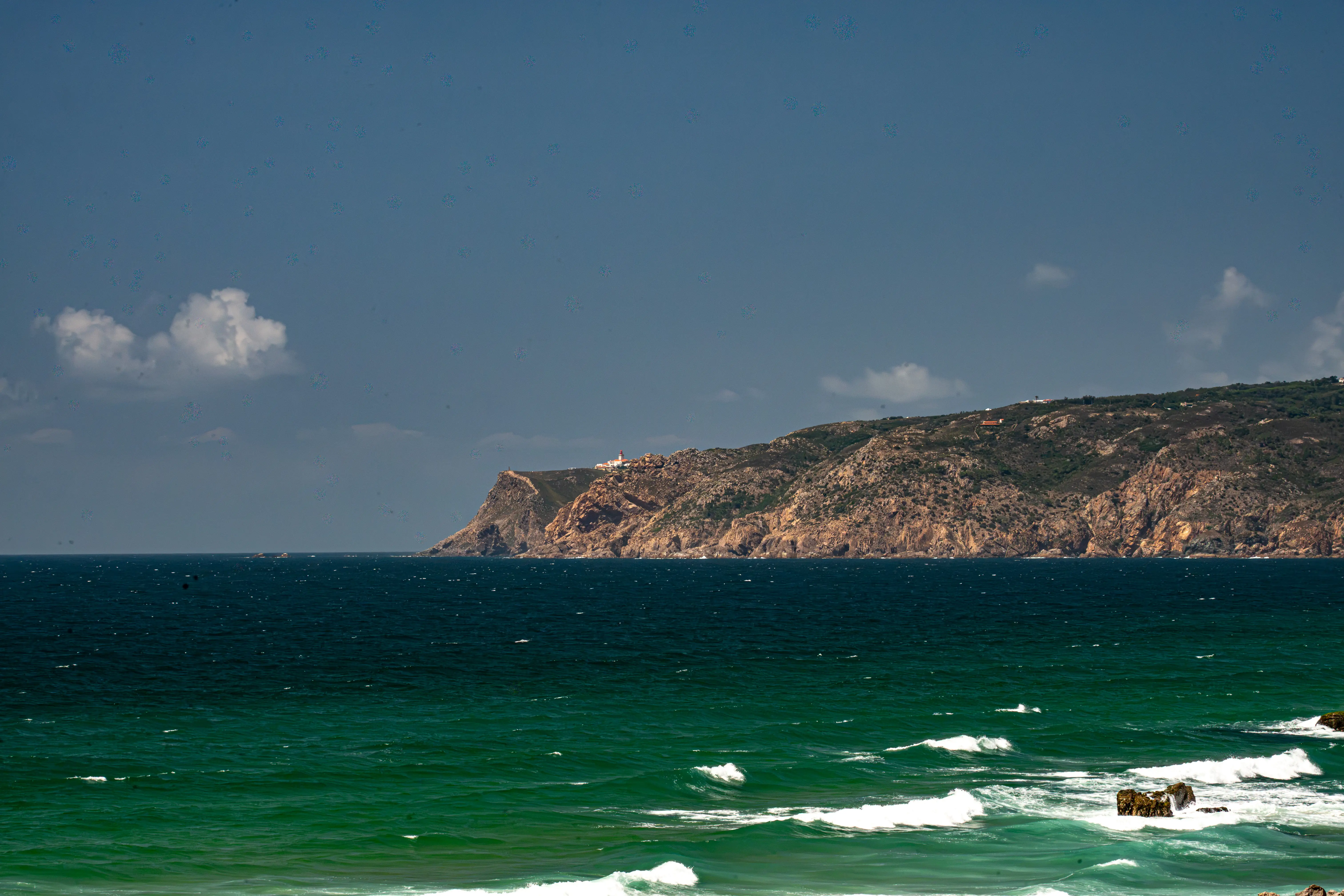Wide shot of a dark, brooding sky over a rugged, brown and green cliff face that meets a deep green ocean. Small white waves are crashing on the shore and around rocks. Buildings are visible on the clifftop in the distance.