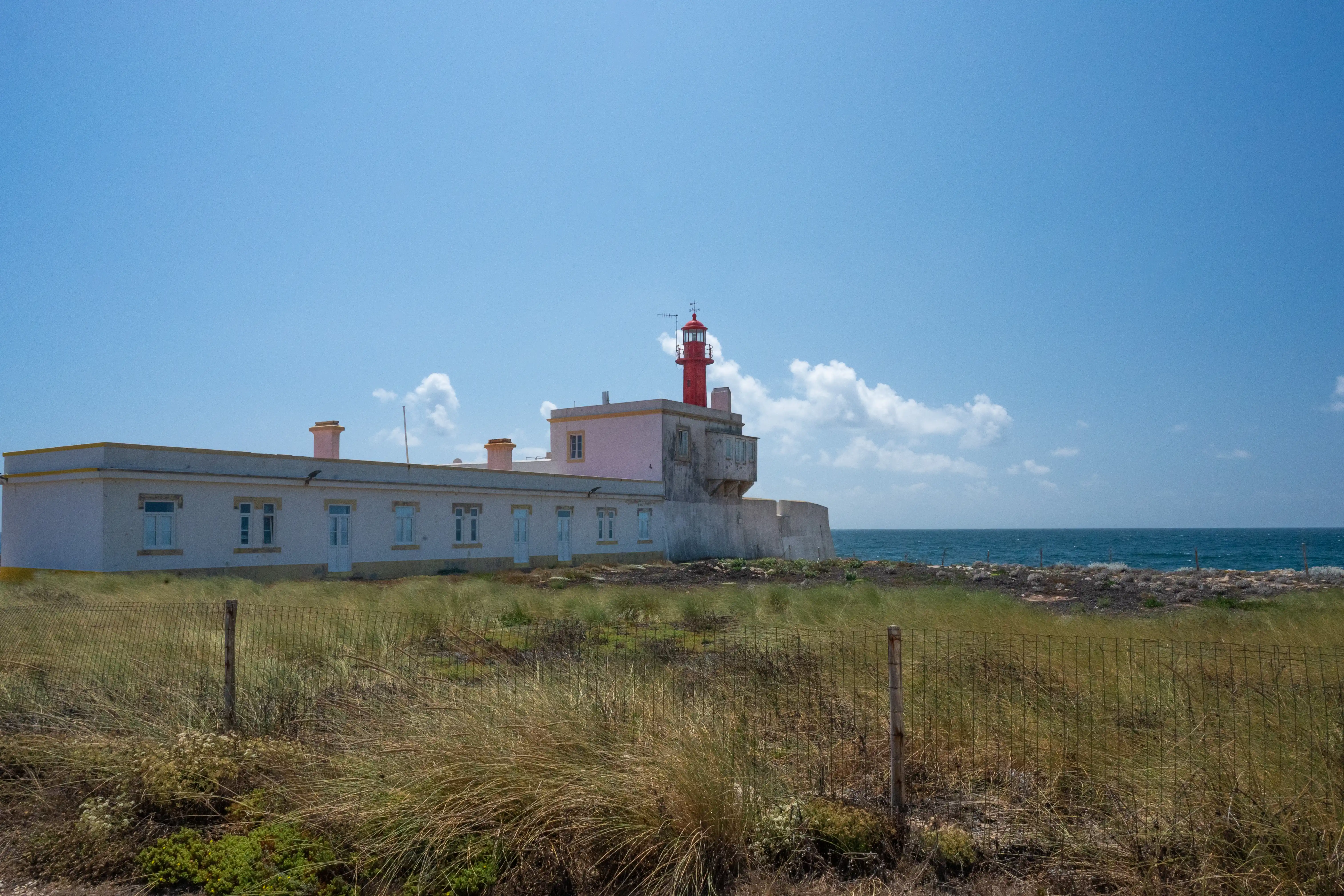 A red lighthouse is attached to a long, white building with several windows and chimneys, set against a bright blue sky with some white clouds. The building is bordered by a low, grey wall. In front of the building, dry, tall grasses are visible, separated from the viewer by a rustic wire fence. The dark blue ocean stretches out to the right.