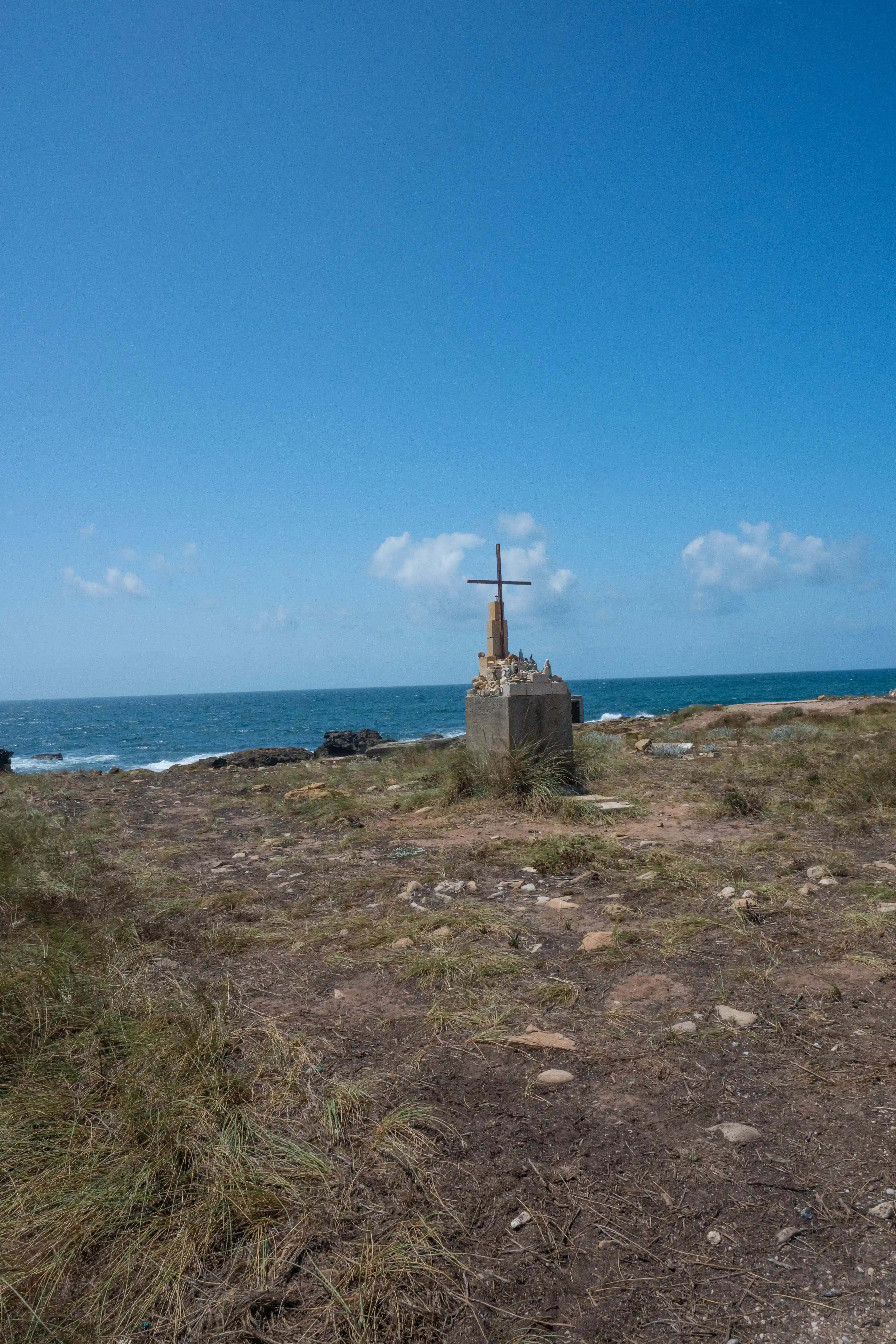 A cross on a concrete plinth sits on a grassy, rocky coastline with the sea and sky in the background.