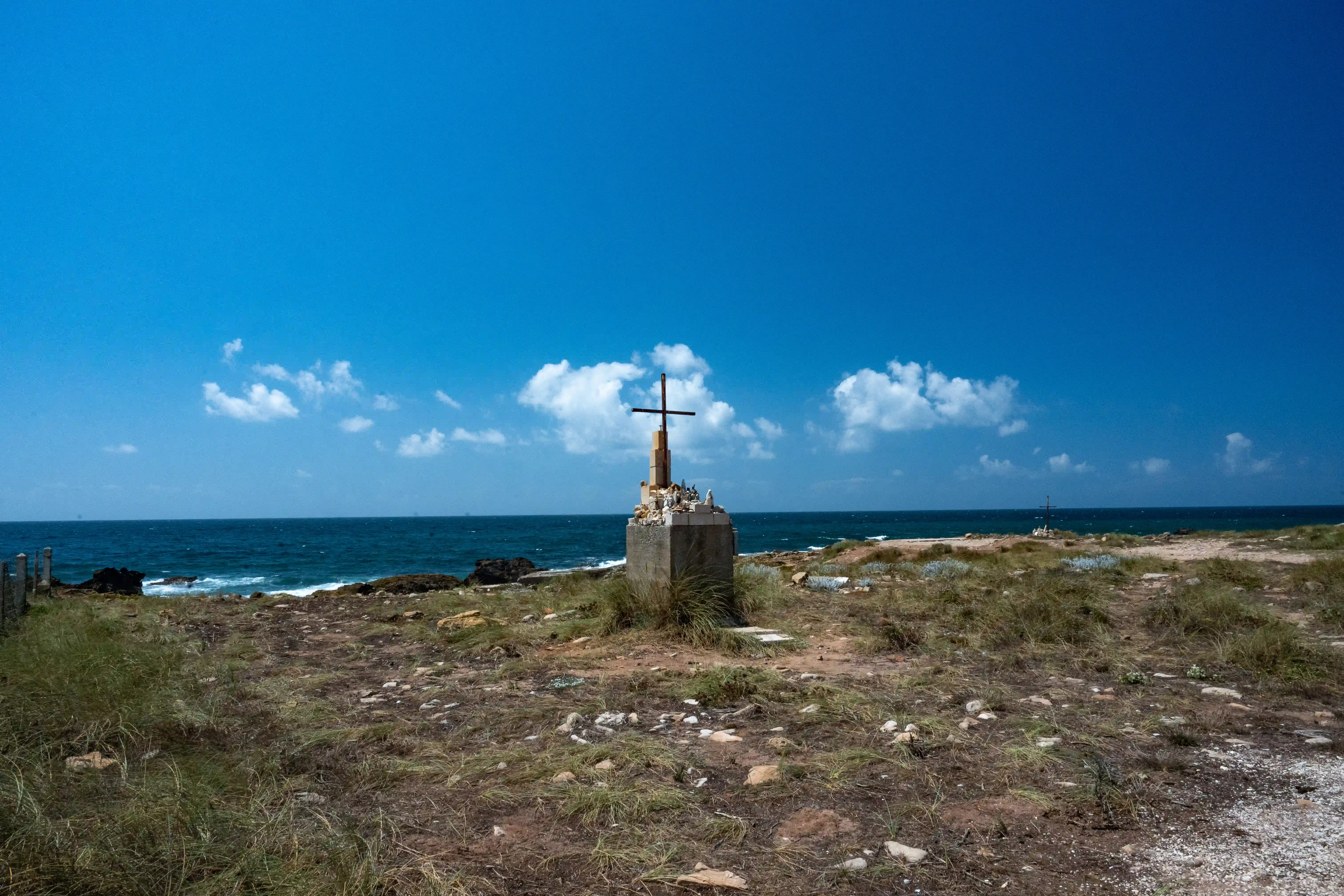 A stone cross monument on a desolate coastal landscape with the sea and a blue sky in the background.