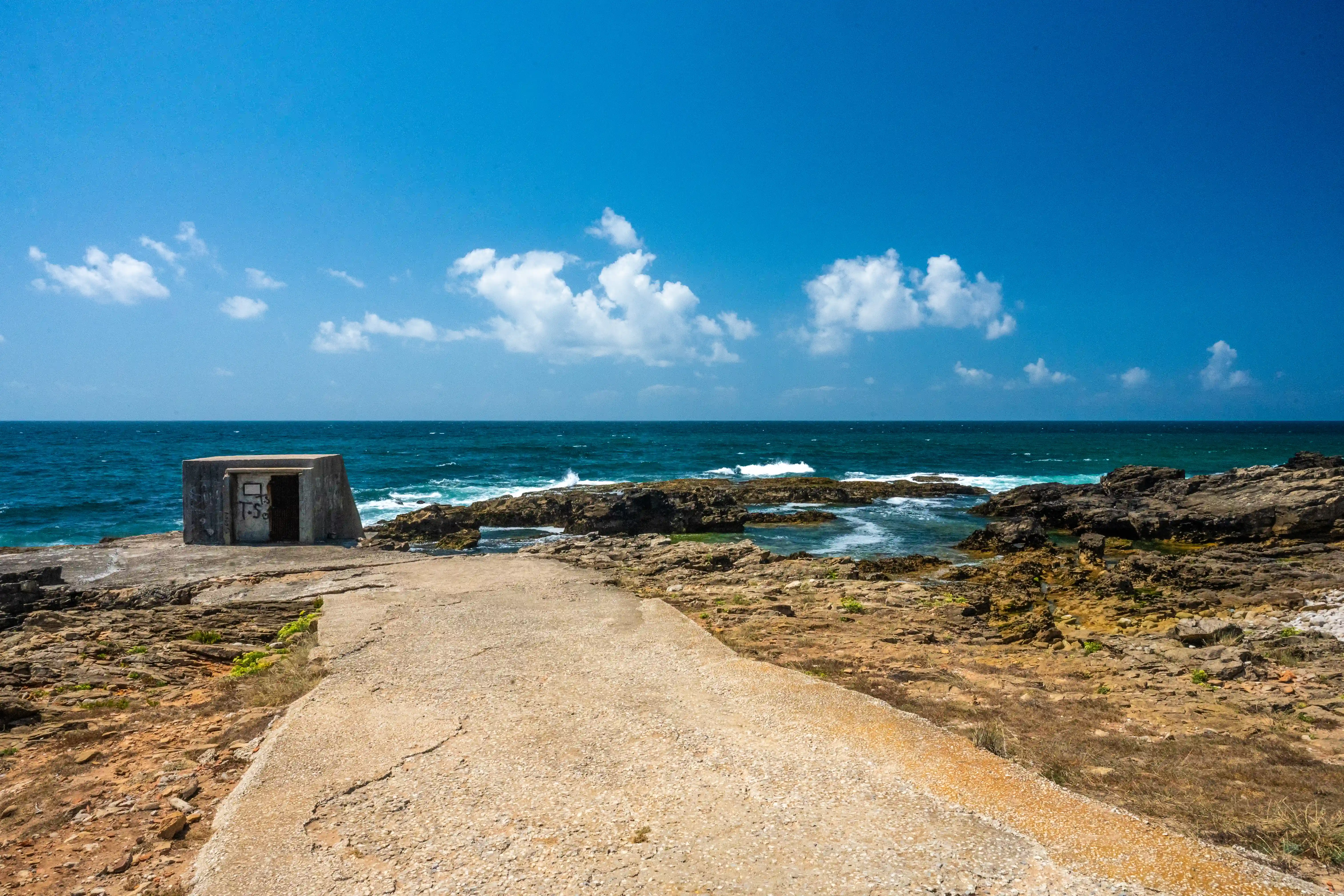 A concrete structure with graffiti stands on a rocky coast, facing a dark, wavy ocean under a blue sky with scattered clouds. A concrete path leads towards it from the foreground.