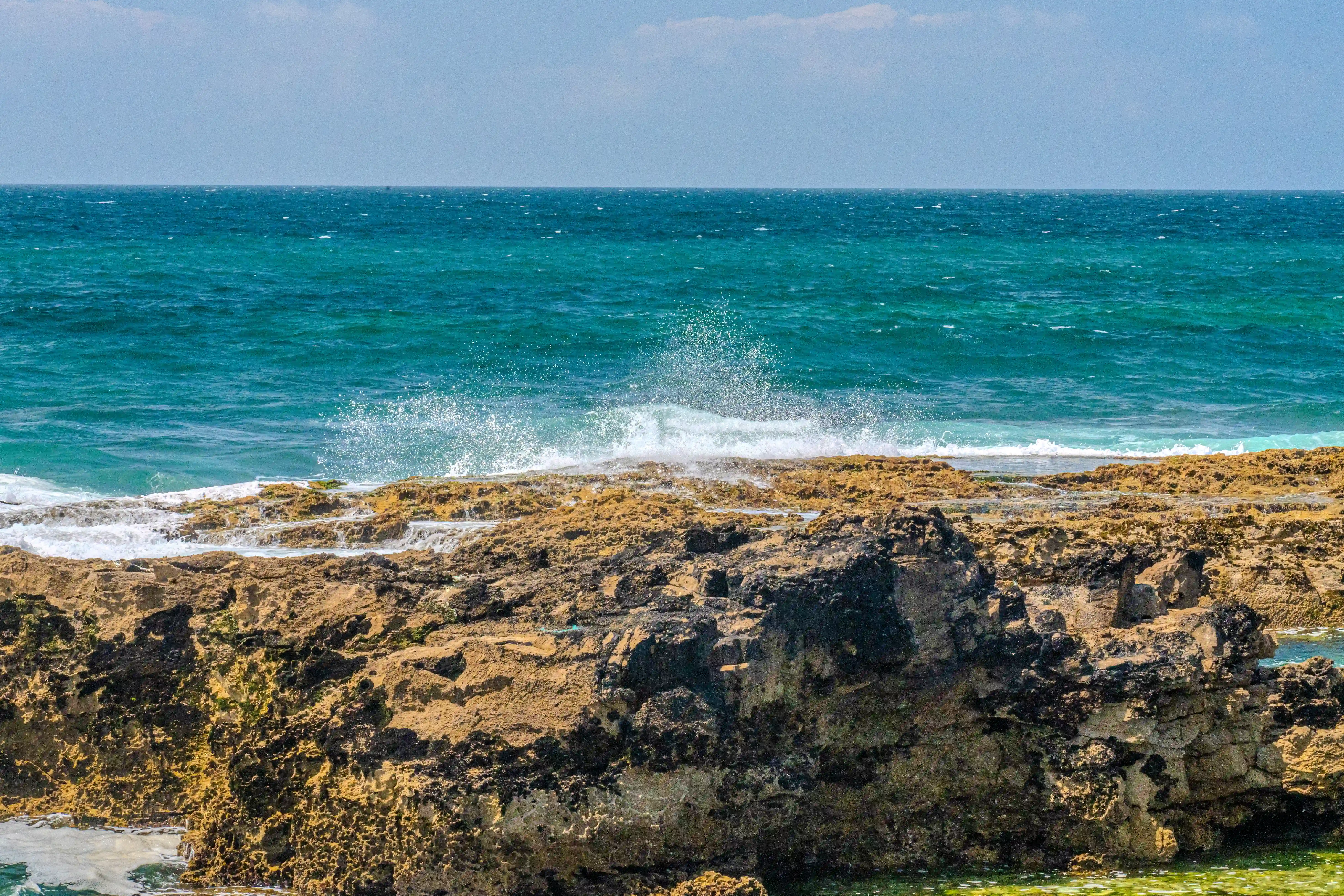 A seascape showing dark, choppy ocean waves breaking against a rough, dark brown rocky shore under a cloudy grey sky.