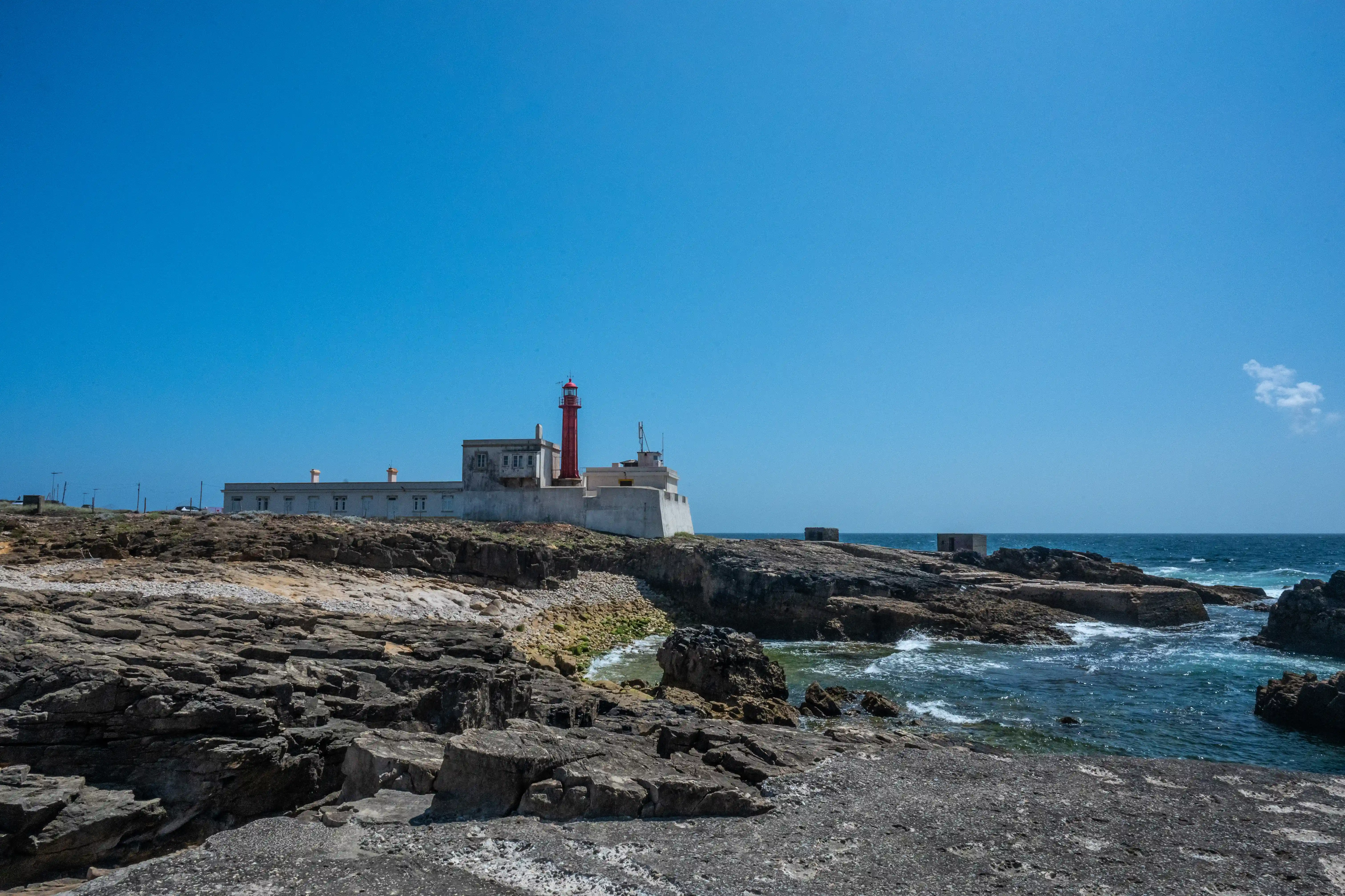 A red lighthouse and white buildings are positioned on a cliff overlooking the sea, with a clear blue sky above. The foreground is made up of dark rocks and pebbles.