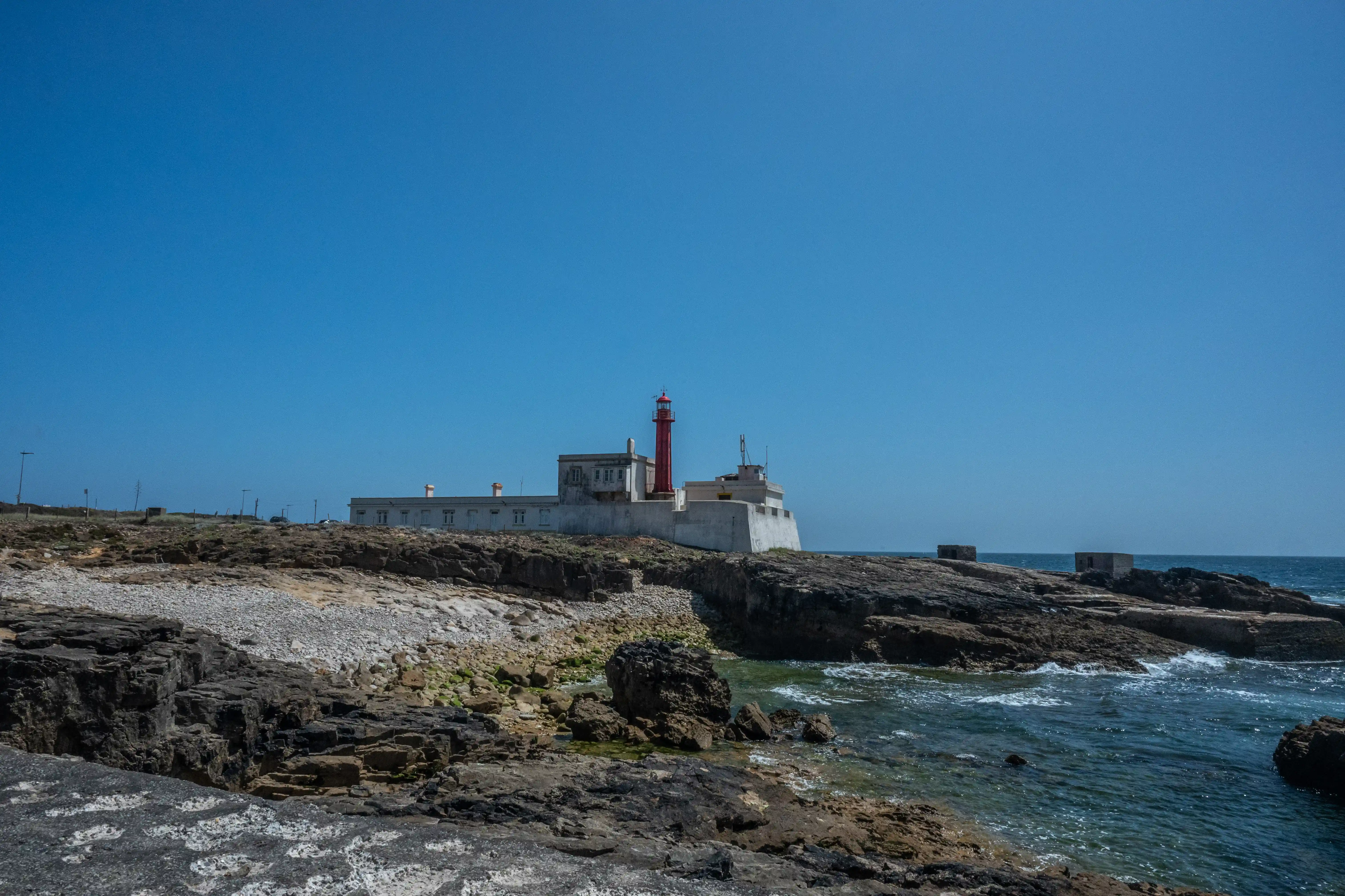  A red lighthouse and white buildings are positioned on a cliff overlooking the sea, with a clear blue sky above. The foreground is made up of dark rocks and pebbles.