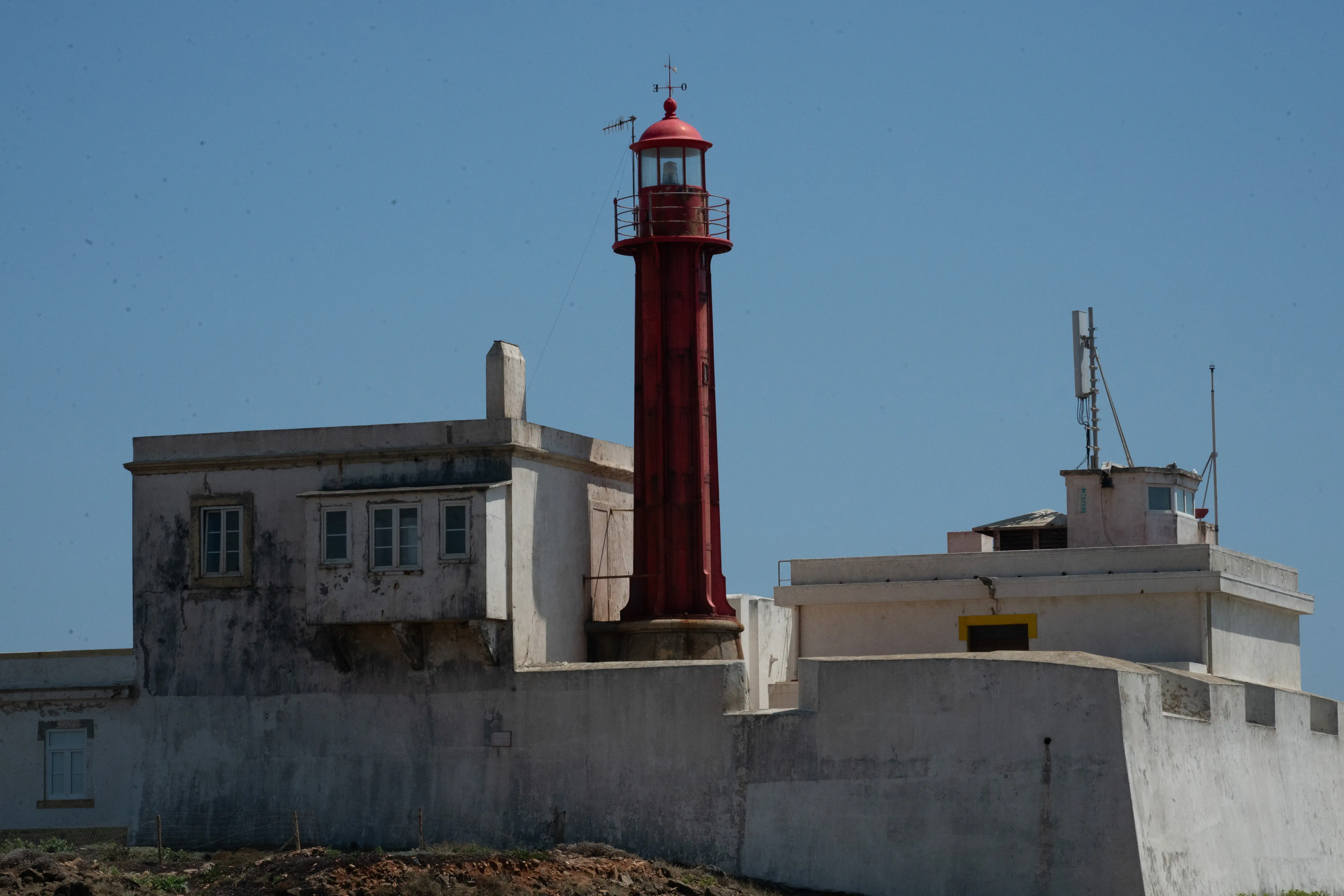 A tall, red lighthouse with a lantern room and railing stands next to a low, white building with windows and a chimney. A modern antenna is visible on another building in the background, all under a clear blue sky.