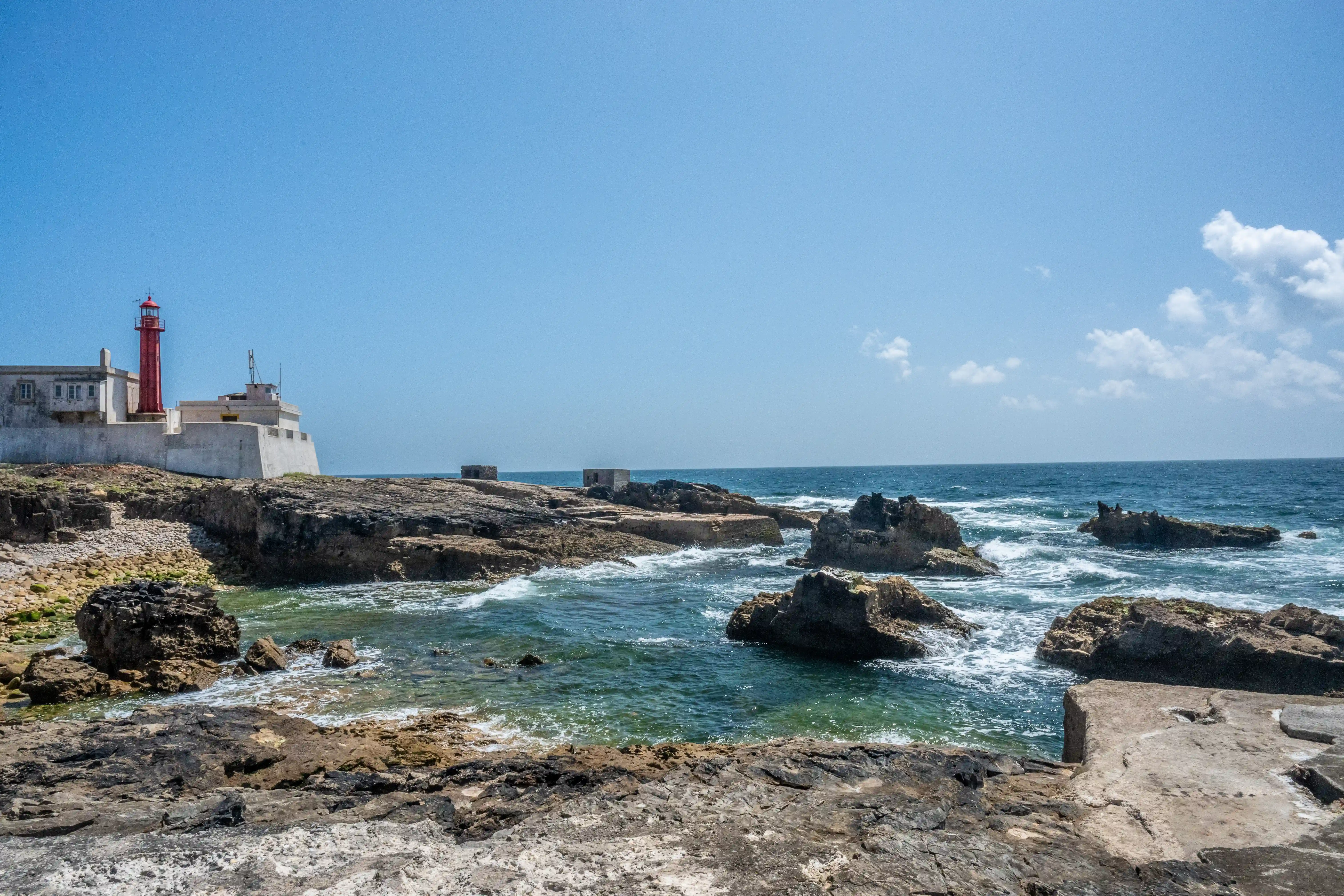 A red and white lighthouse structure on a rocky coast with waves breaking against the shore.