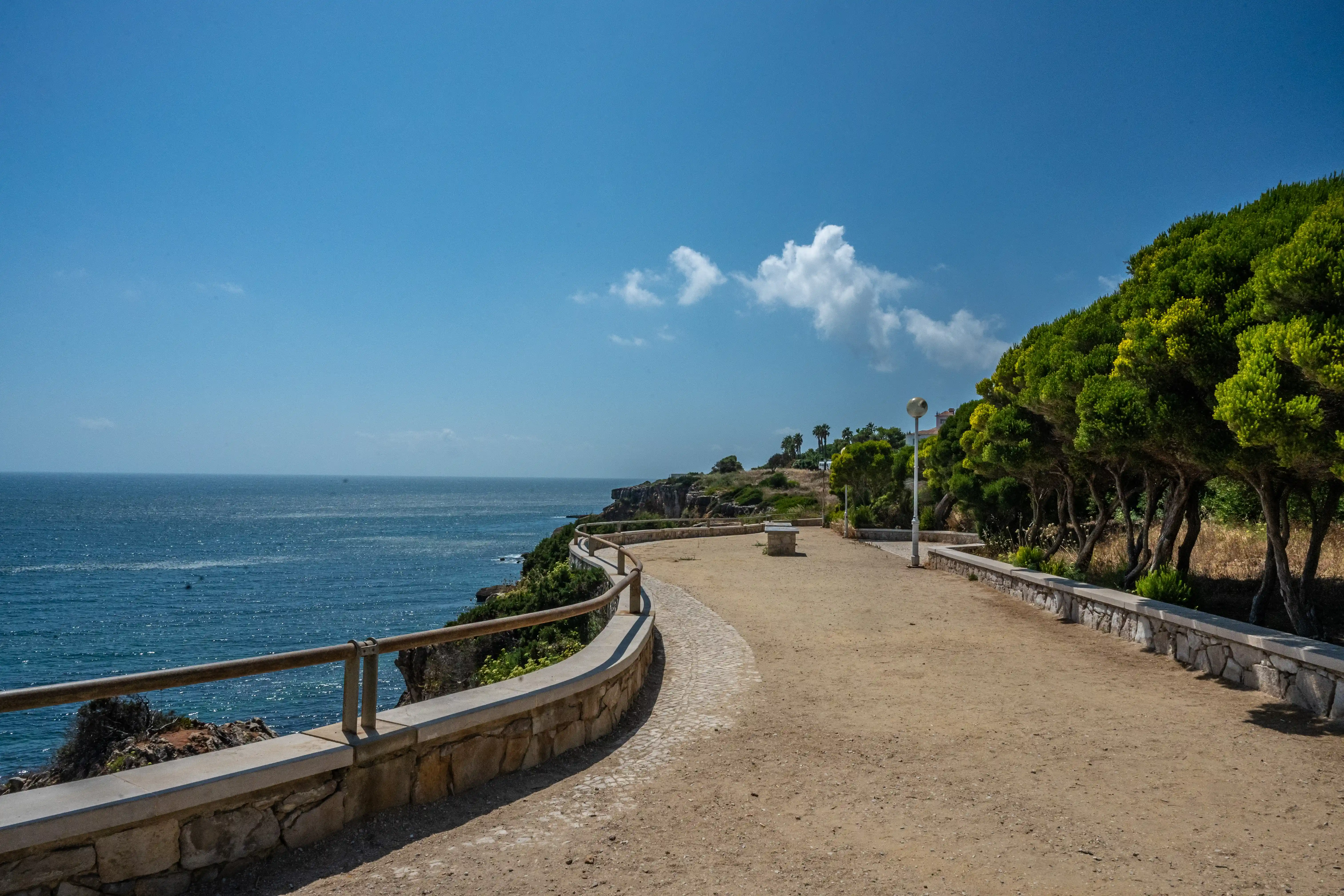  A wide, sandy and paved coastal path with a stone wall and wooden railing curves to the left, overlooking a calm blue sea. Green trees line the right side of the path, and palm trees are visible on a hill in the distance under a bright blue sky with scattered white clouds.