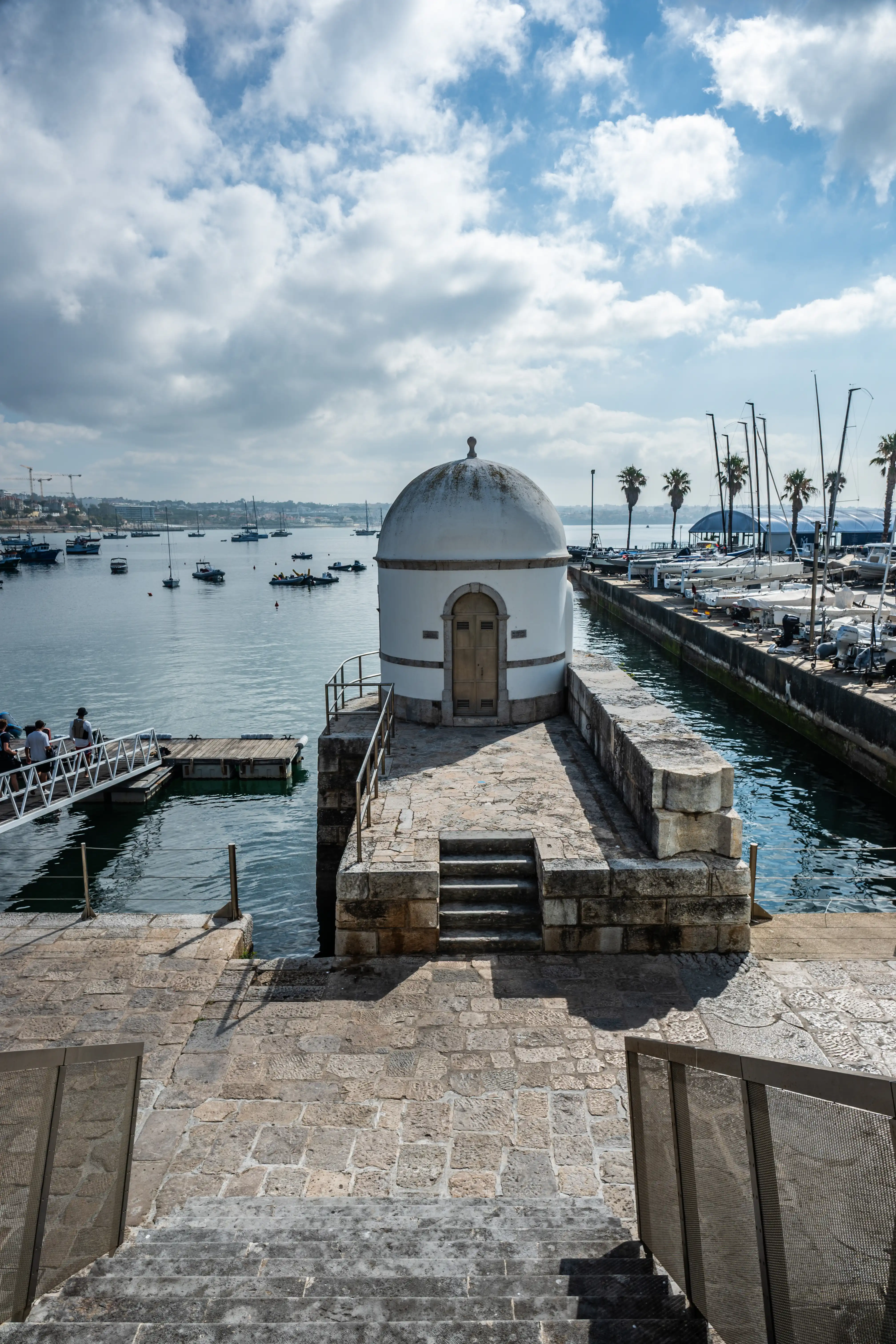 A small, white, cylindrical building with a white dome and a pale wooden door is situated on a stone pier. The pier has stone steps leading down to the water and metal railings. In the background, a harbour is visible with various boats, including sailboats and smaller dinghies, moored in the water. Palm trees and distant buildings can be seen on the shoreline under a bright, cloudy sky.