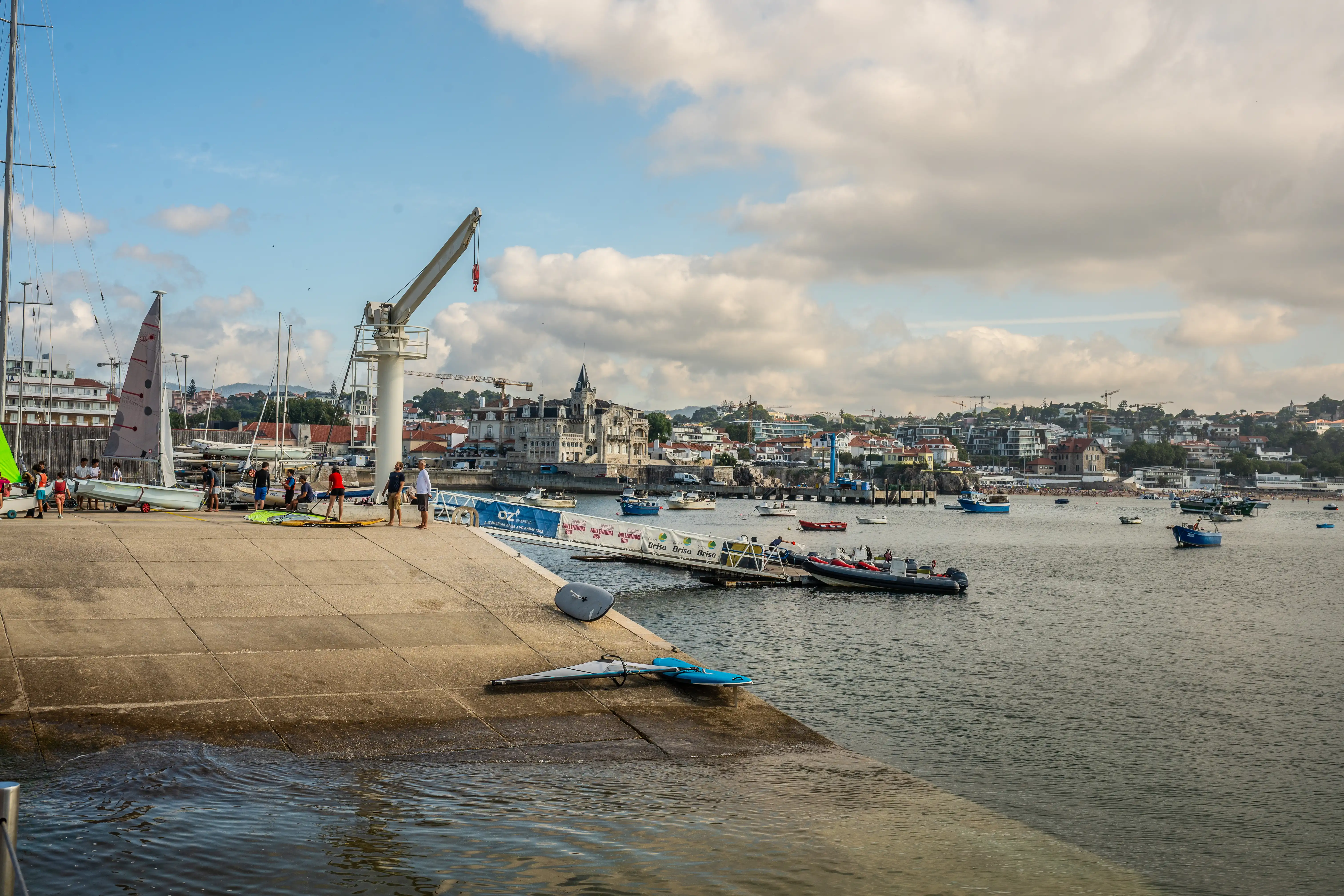 People are gathered on a concrete pier next to several sailboats, some on trailers. In the water, various boats are moored, and a ramp leads to a floating pontoon where a few people stand. A crane is positioned on the pier. Beyond the harbour, a scenic town with distinctive architecture stretches along the coastline, backed by hills