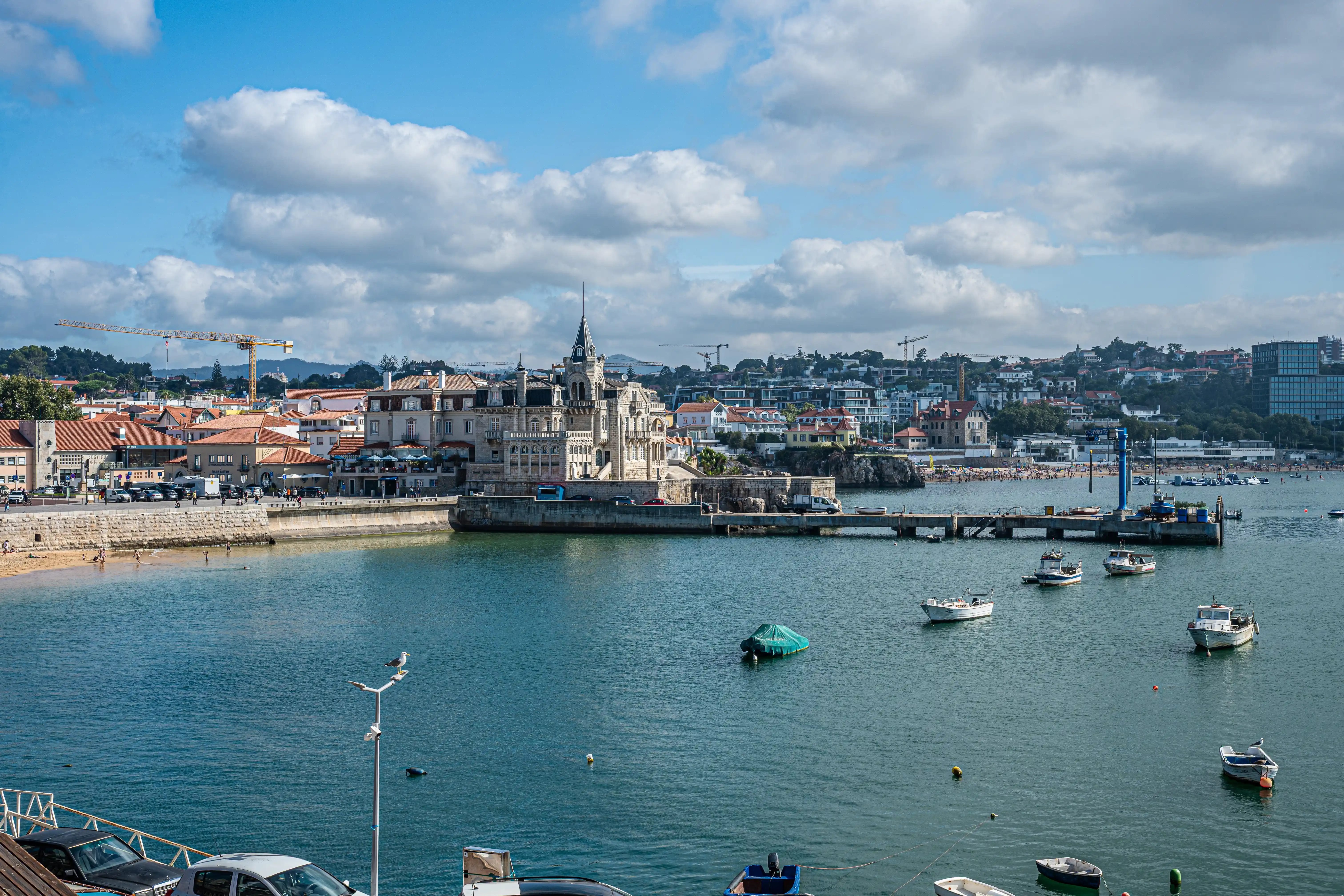 A wide shot of a coastal town with a grand building on the waterfront, a harbour filled with boats, and a beach in the background.