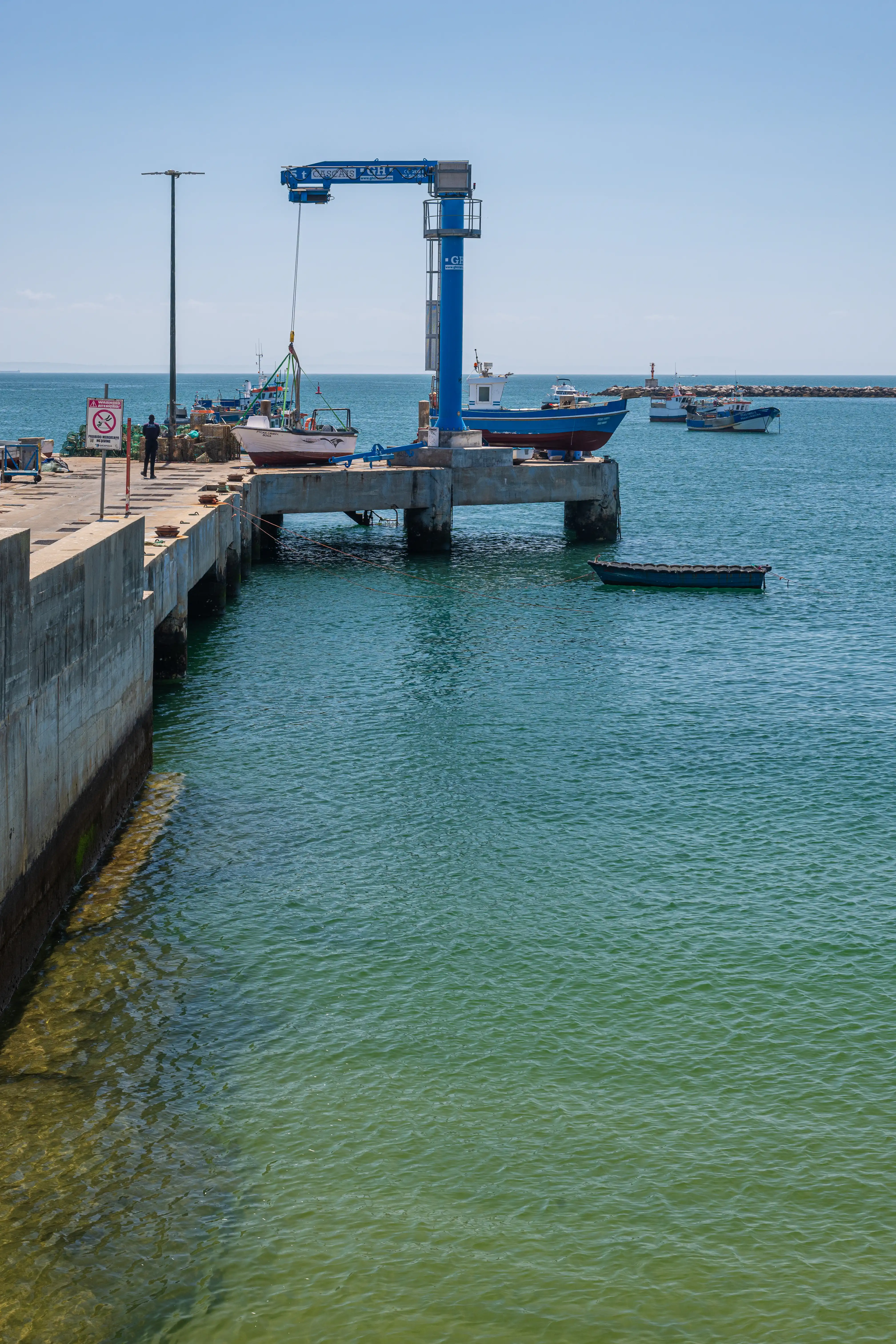 A blue gantry crane is lifting a white boat out of the water at a harbour. There are several other boats in the sea.