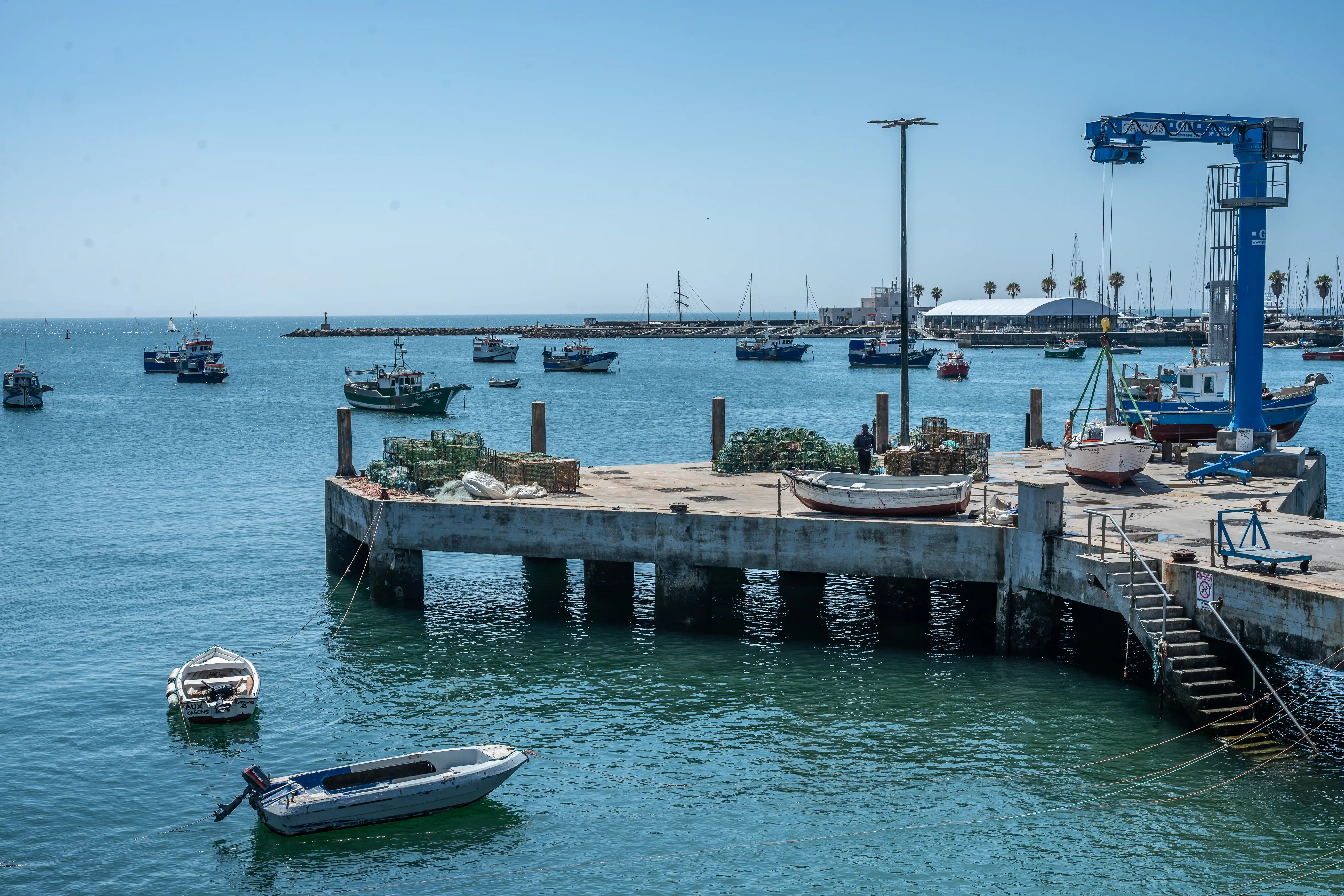 A busy harbour with many fishing boats, a concrete pier with fishing gear, and a large blue crane lifting a small sailboat.