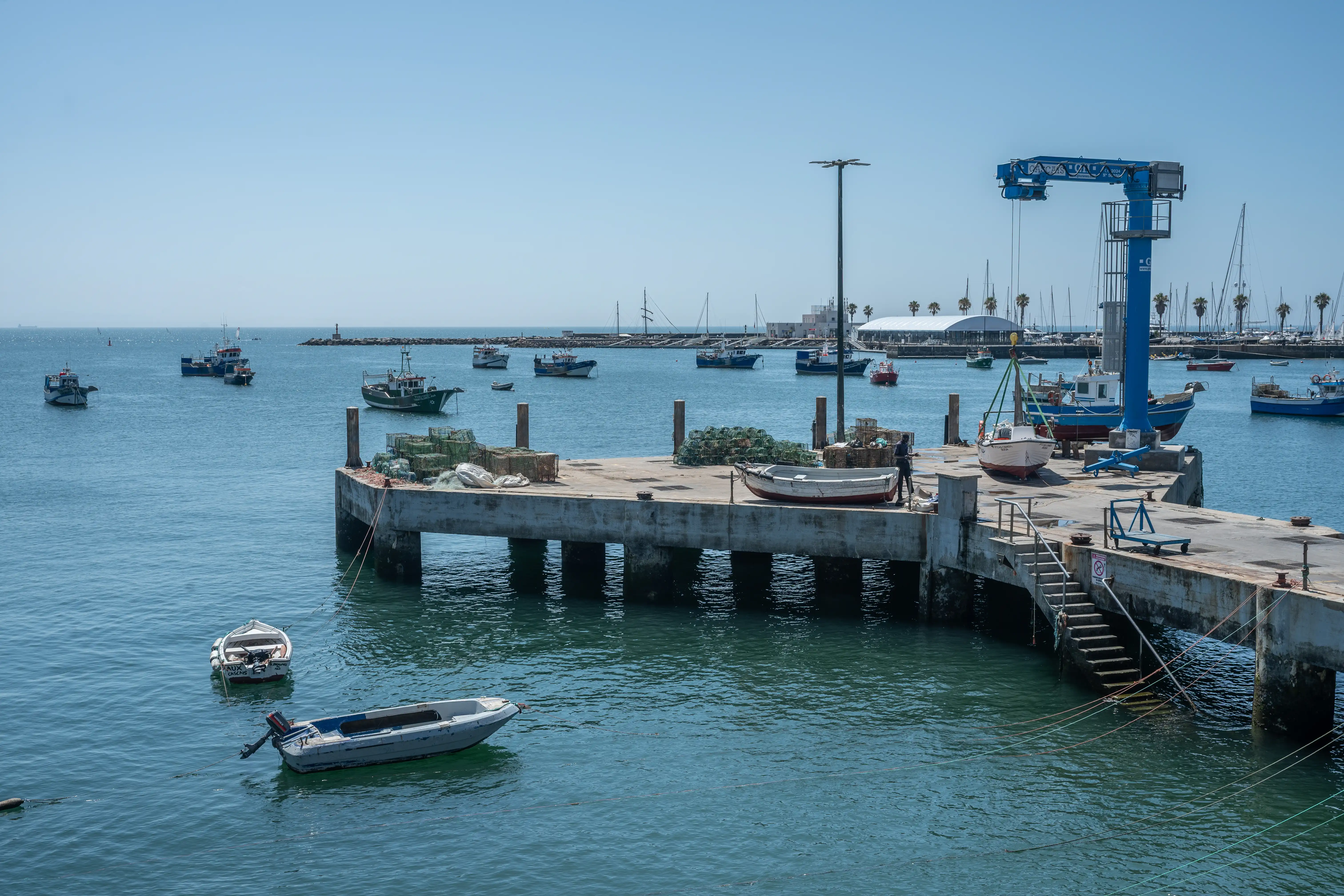 A busy harbour with many fishing boats, a concrete pier with fishing gear, and a large blue crane lifting a small sailboat.