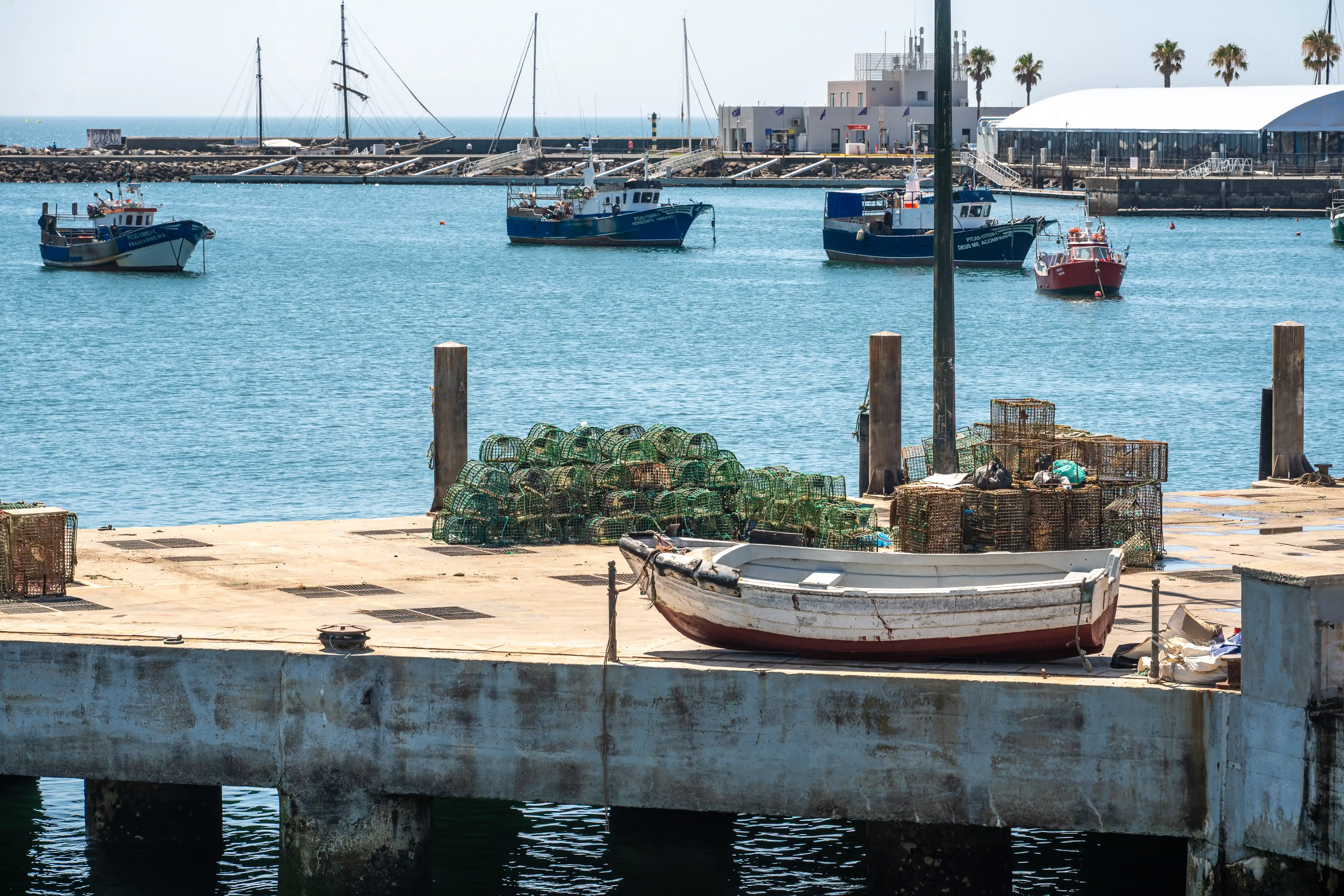 A harbour scene showing several fishing boats in the blue water, with a concrete dock in the foreground where many green lobster pots are piled up and a small white rowing boat is tied.