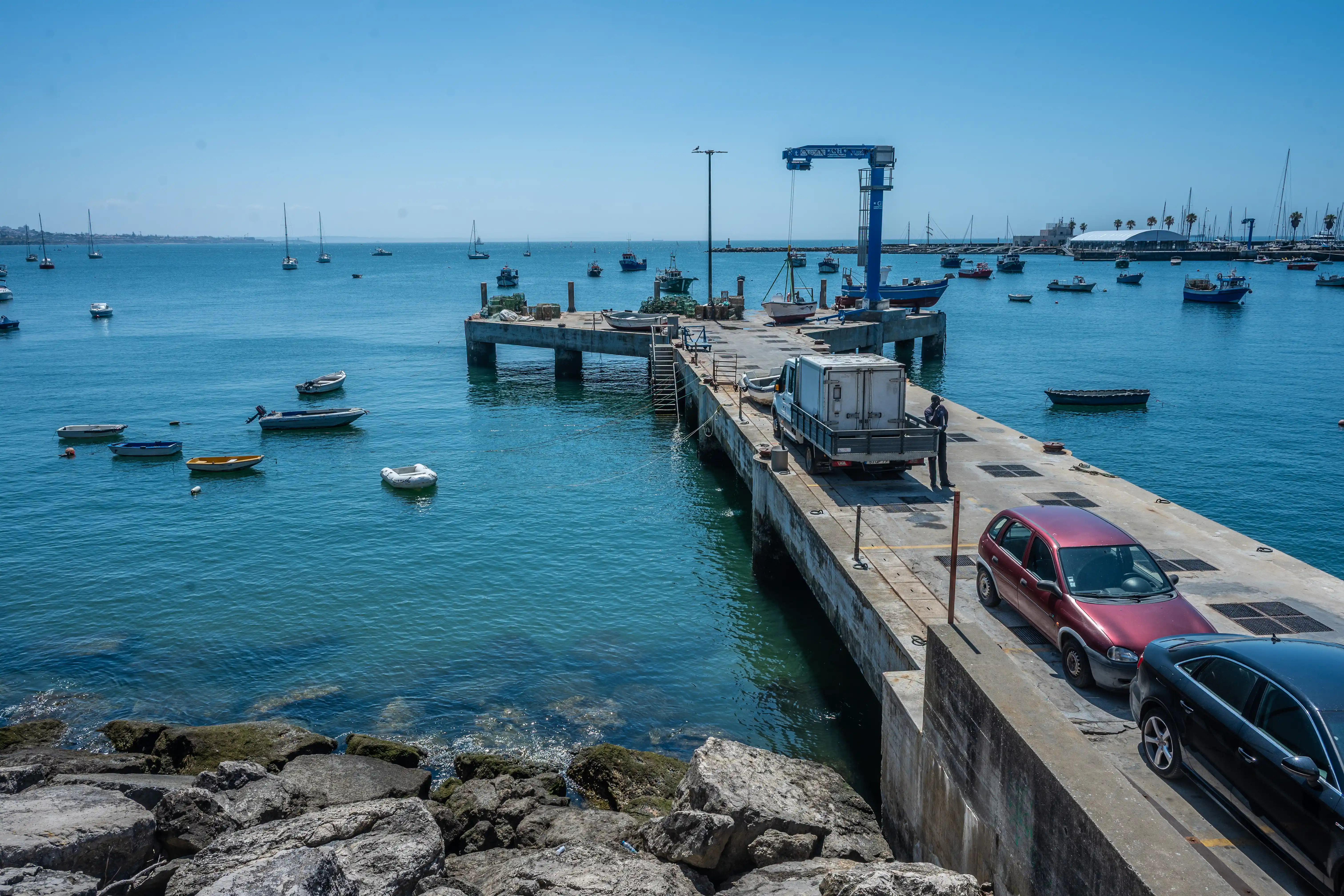 Coastal pier with small boats, parked cars, a crane structure, and a marina in the background.