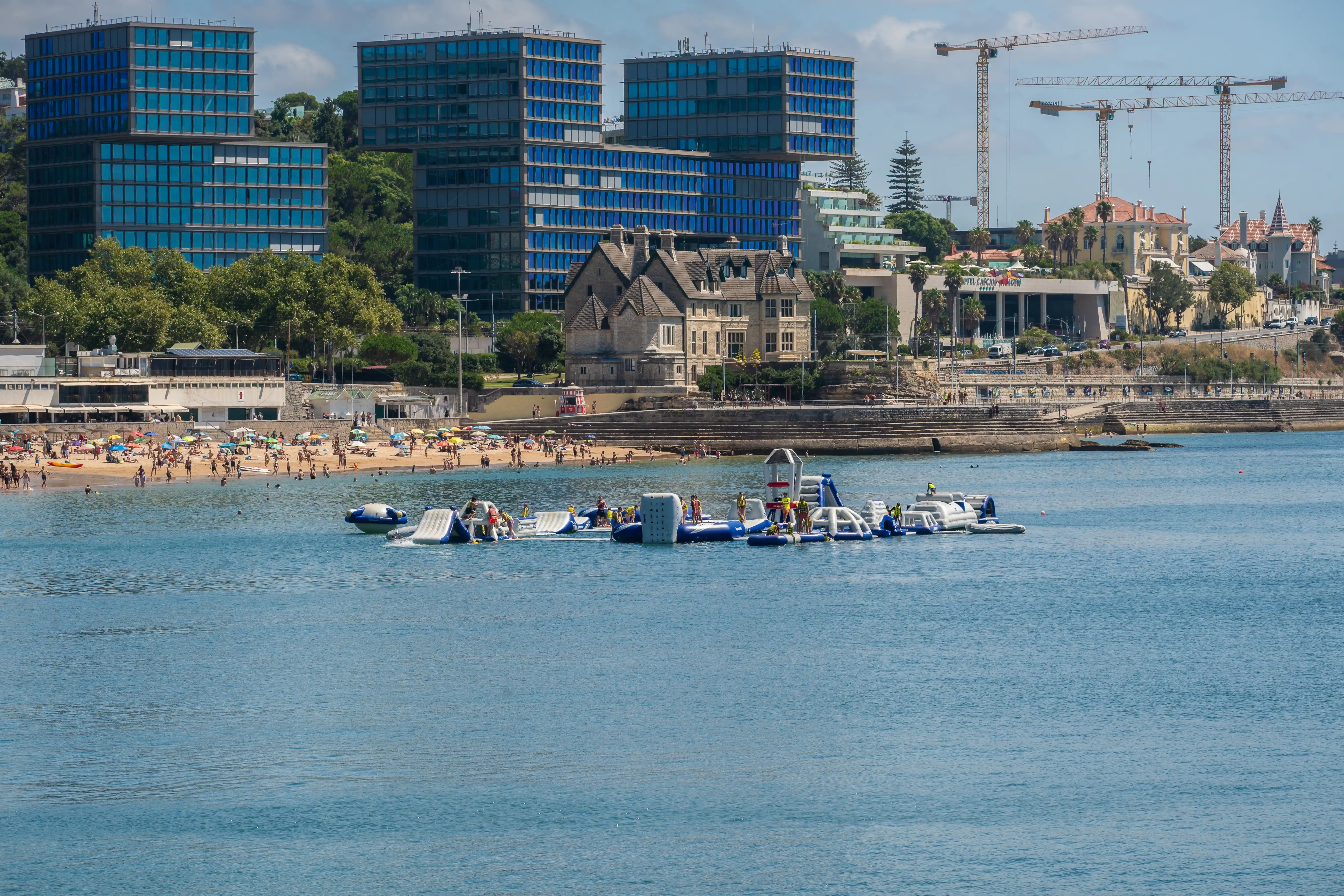 Busy beach with swimmers, an inflatable water park, and modern buildings with cranes in the background.