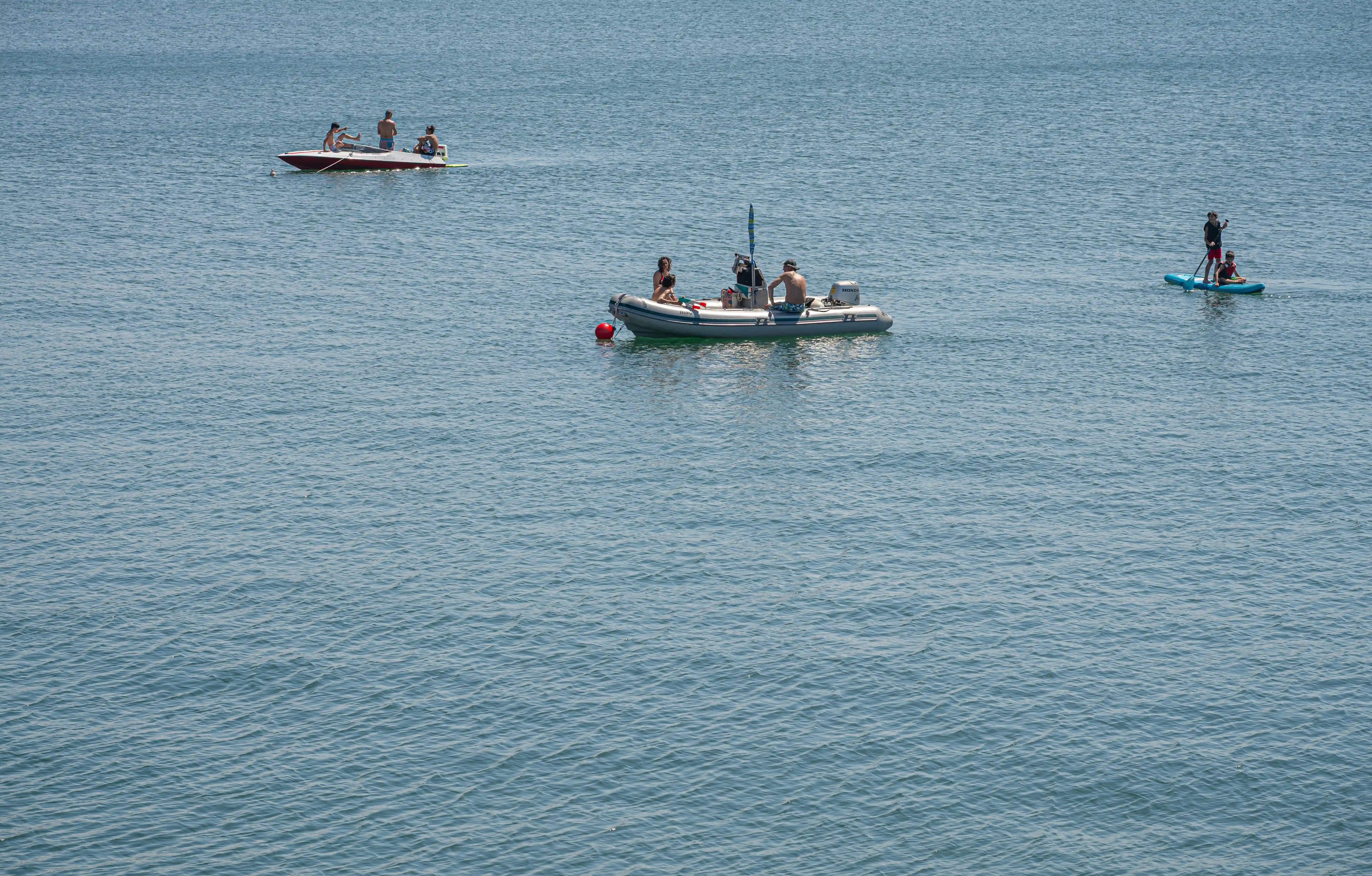 Several people are enjoying various watercraft on a blue, rippling body of water. A RIB boat with three people is in the centre, a person on a paddleboard with another seated is to the right, and a speed boat with three people is in the mid-left.