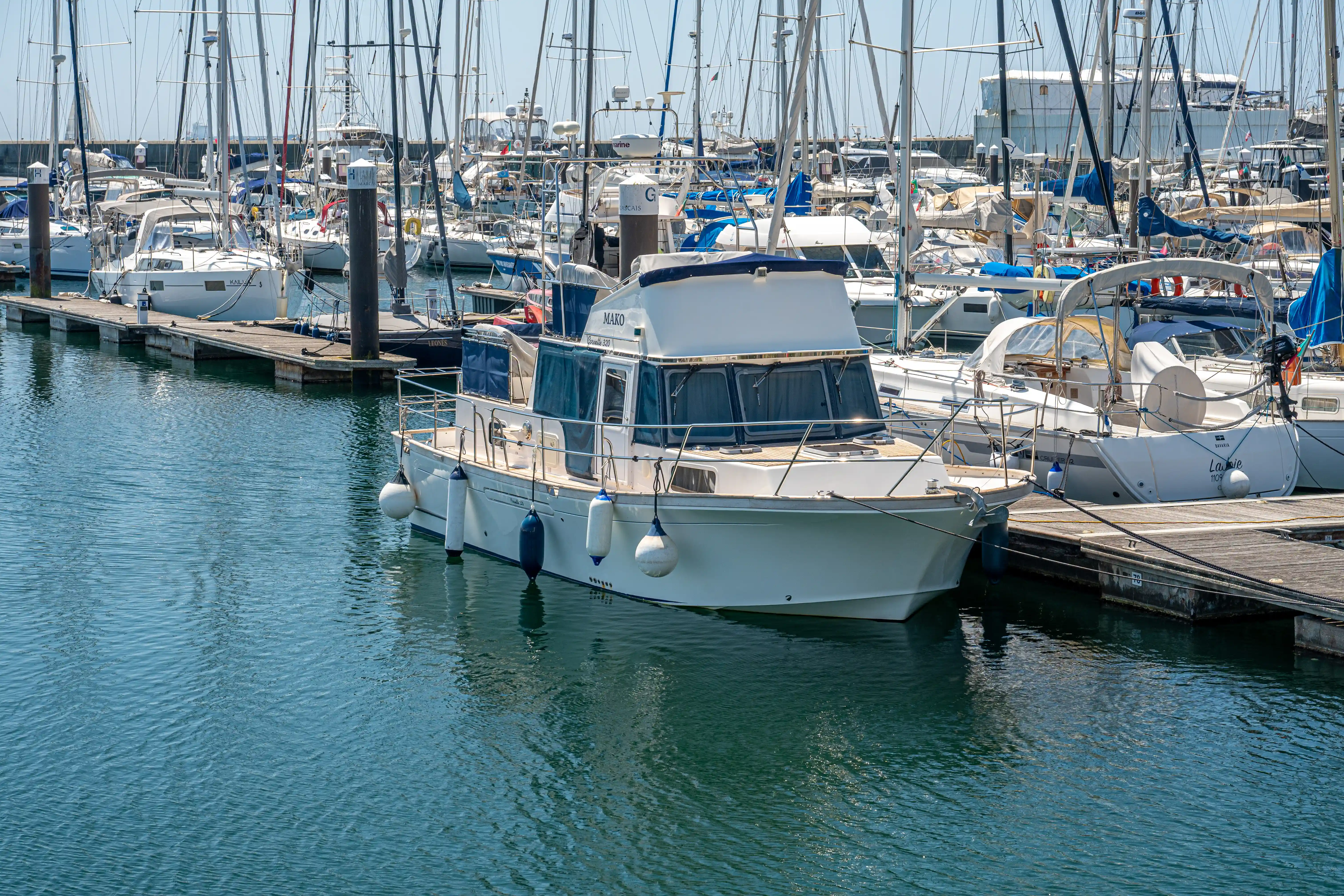 A white motorboat named 'MAKO' is docked in a marina next to a wooden pier. Numerous sailboats with tall masts are moored in the background. The water is blue and calm, reflecting the sunny sky and the boats.