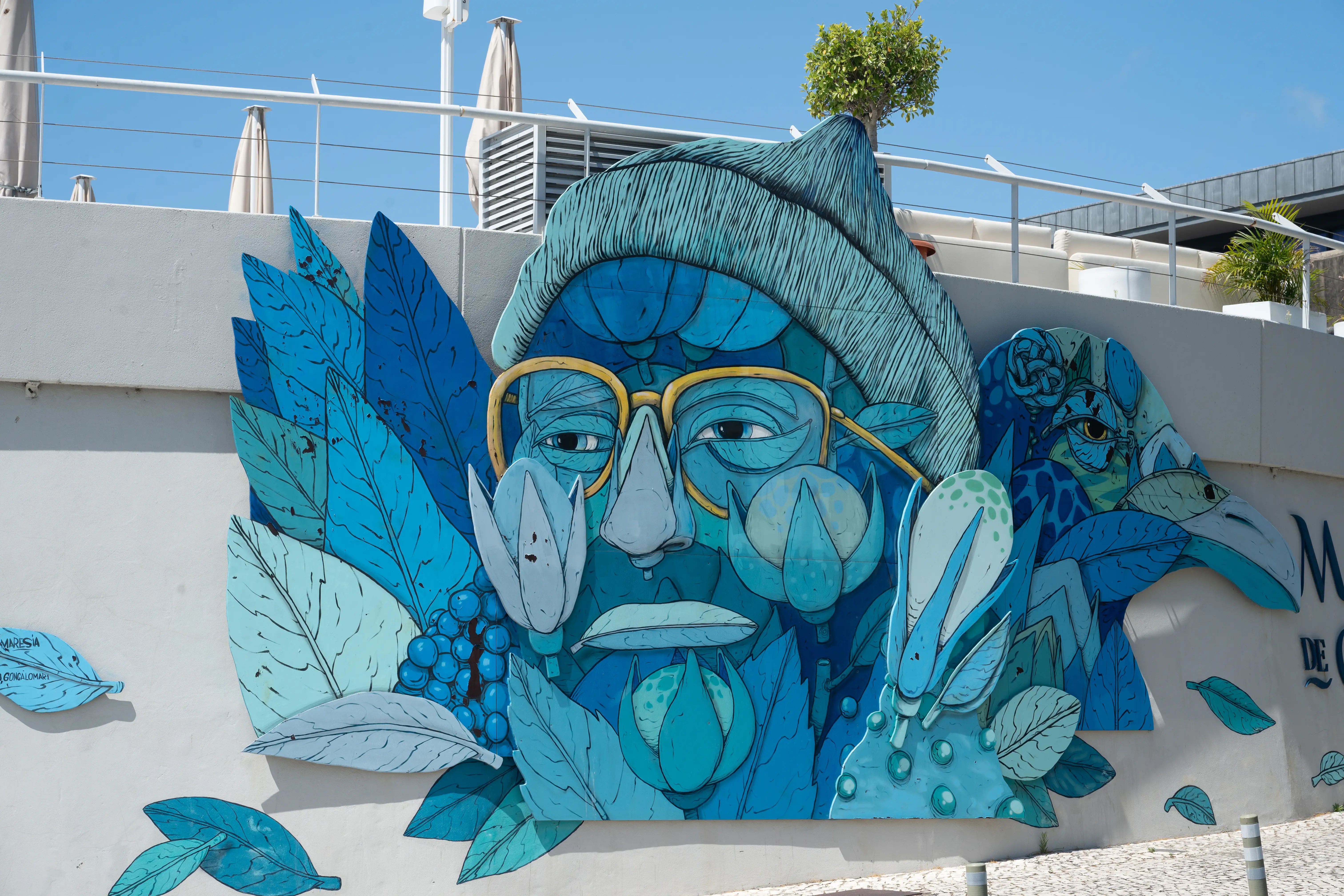 A large, colourful mural painted on a white wall features a face rendered in various shades of blue, with yellow-rimmed glasses. The face is surrounded by abstract shapes resembling leaves and flowers, also in blue hues.