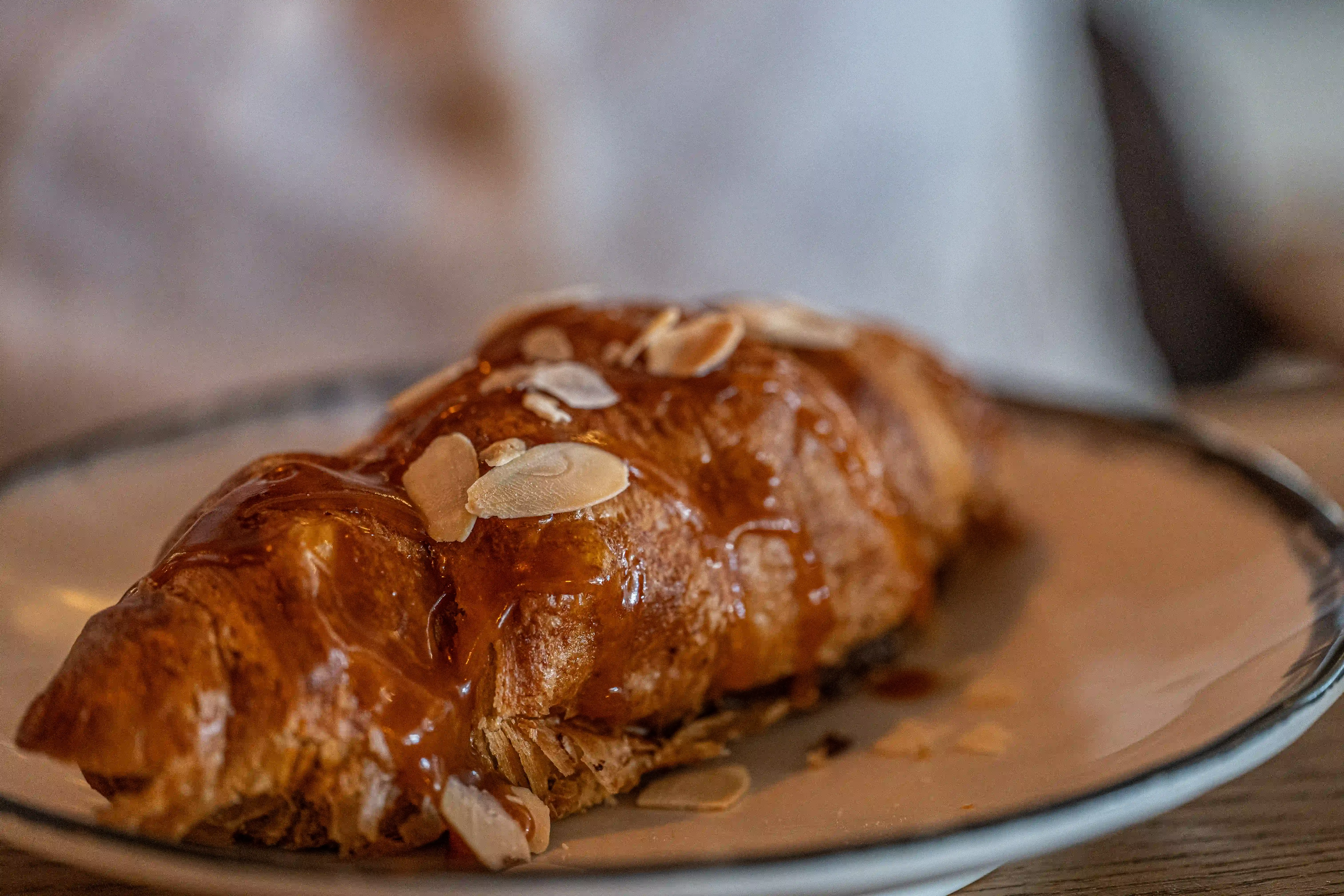 A baked croissant, topped with a shiny caramel glaze and sliced almonds, sits on a plate.