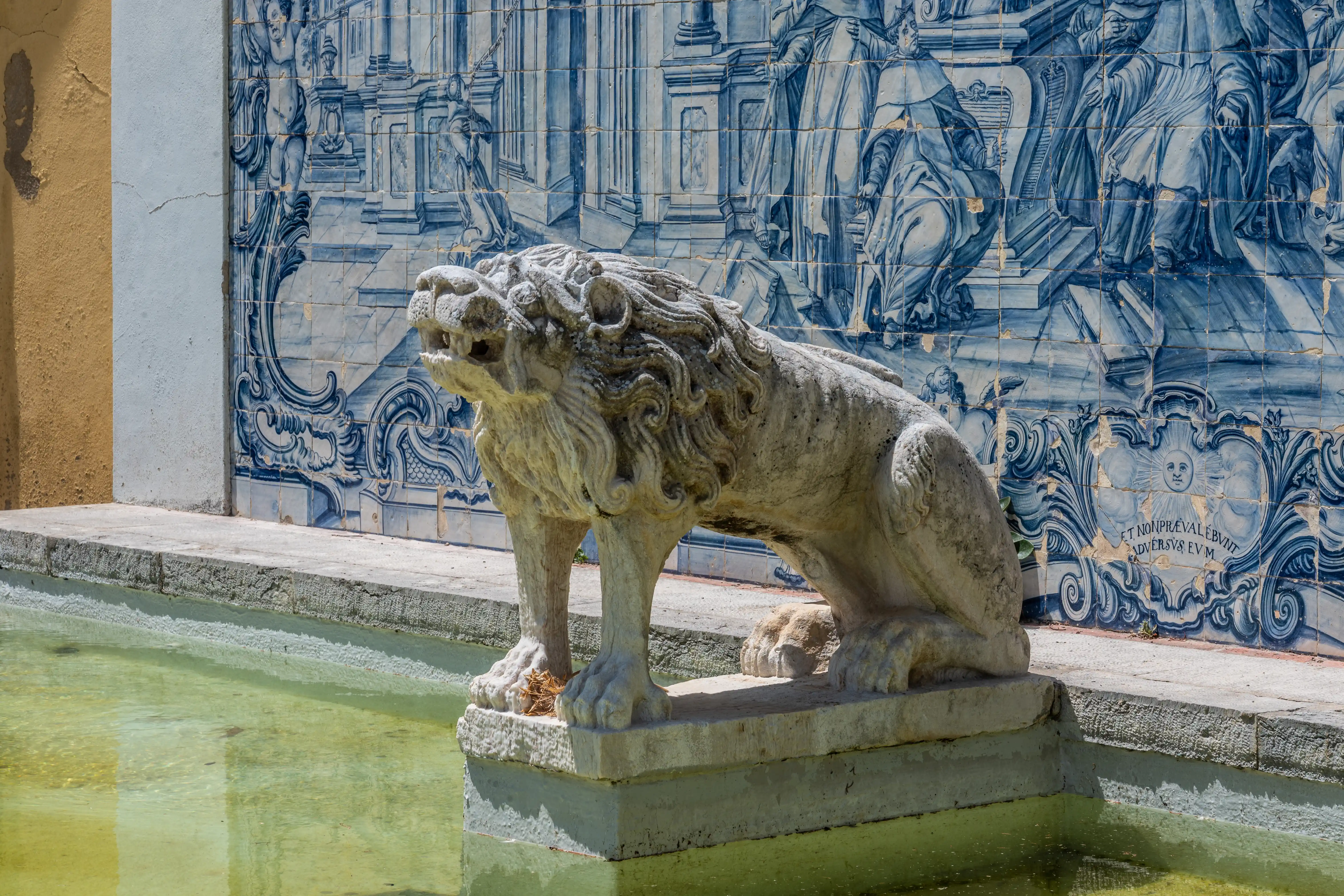  A stone lion sculpture with its mouth open sits on a pedestal next to a pool of water. Behind the statue is a wall decorated with blue and white historical azulejo tiles featuring figures and ornate patterns.