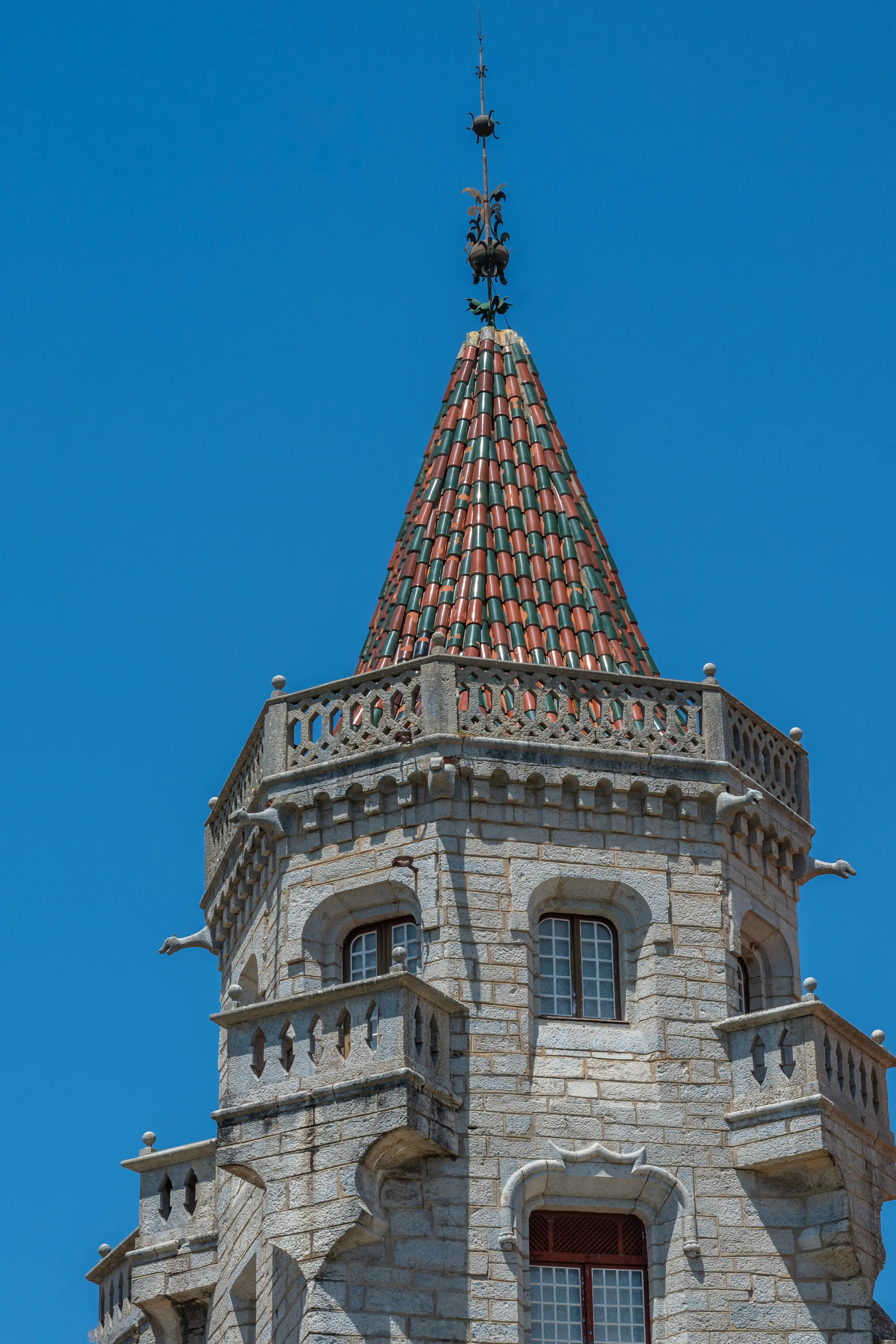 Close-up of a stone tower turret with a red and green tiled conical roof, a decorative metal finial, and intricate stonework, set against a bright blue sky.