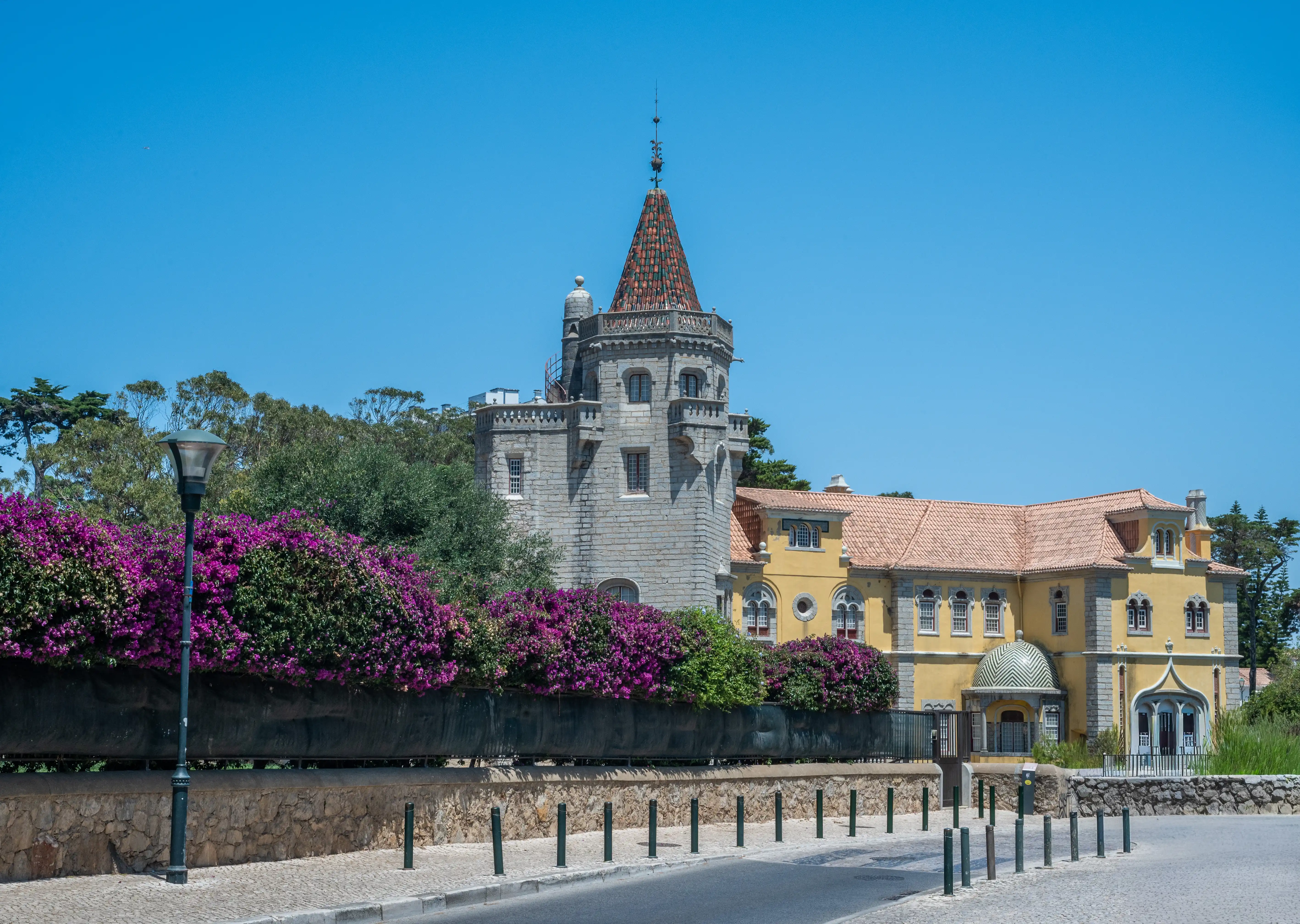 A stone tower with a red-tiled conical roof stands to the left of a yellow building with a terracotta-tiled roof. Vibrant purple bougainvillea flowers cascade over a wall in front of the buildings. People are walking on a cobbled path in the foreground.